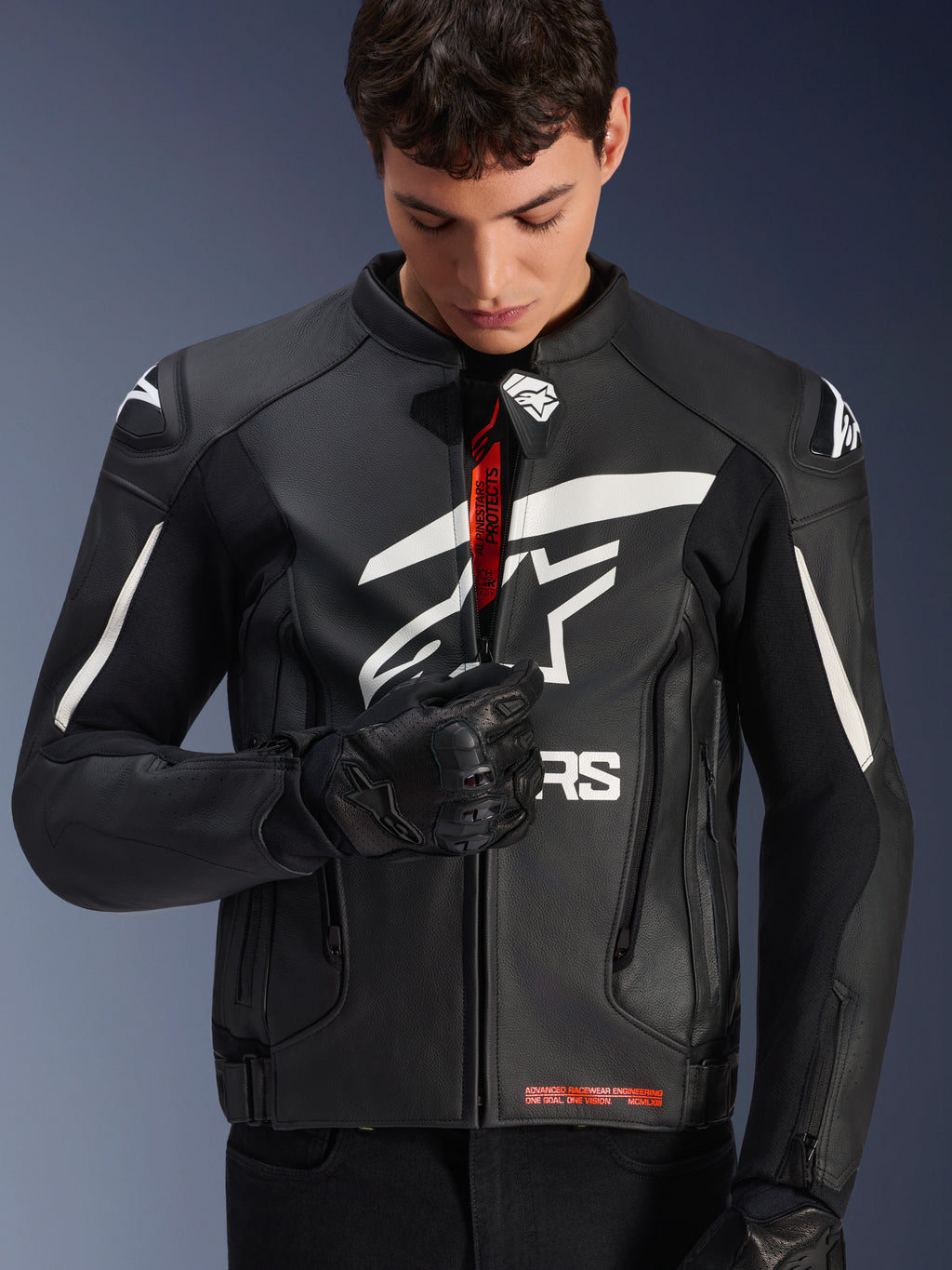 Blouson en cuir Alpinestars GP Plus V4, noir et blanc, modèle masculin fermant la fermeture éclair avant pour révéler le marquage intérieur rouge, avec des logos Alpinestars blancs sur la poitrine et les épaules, porté avec des gants de moto en cuir noir, vêtement de pilotage en cuir dérivé de la course