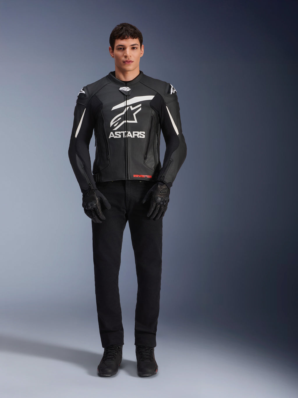 Modèle masculin portant le blouson en cuir Alpinestars GP Plus V4, noir et blanc, avec logo Astars blanc sur la poitrine et détails blancs sur les épaules, accompagné de gants et d'un pantalon de moto noirs, tenue de moto sportive et élégante