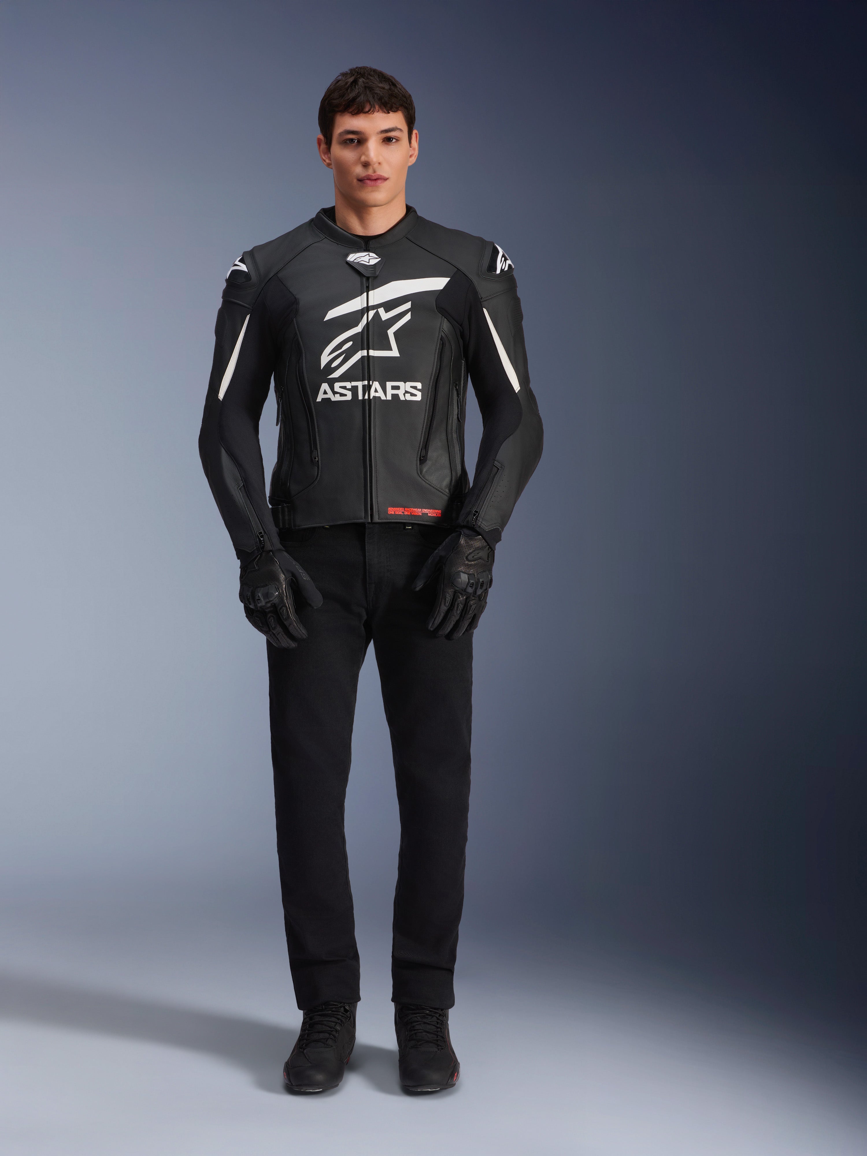 Modèle masculin portant le blouson en cuir Alpinestars GP Plus V4, noir et blanc, avec logo Astars blanc sur la poitrine et détails blancs sur les épaules, accompagné de gants et d'un pantalon de moto noirs, tenue de moto sportive et élégante