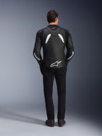 Modèle vu de dos portant le blouson en cuir Alpinestars GP Plus V4 en noir et blanc, associé à un pantalon noir et des bottes de moto, présentant des empiècements blancs sur les épaules et le logo Alpinestars sur les manches et le bas du dos.