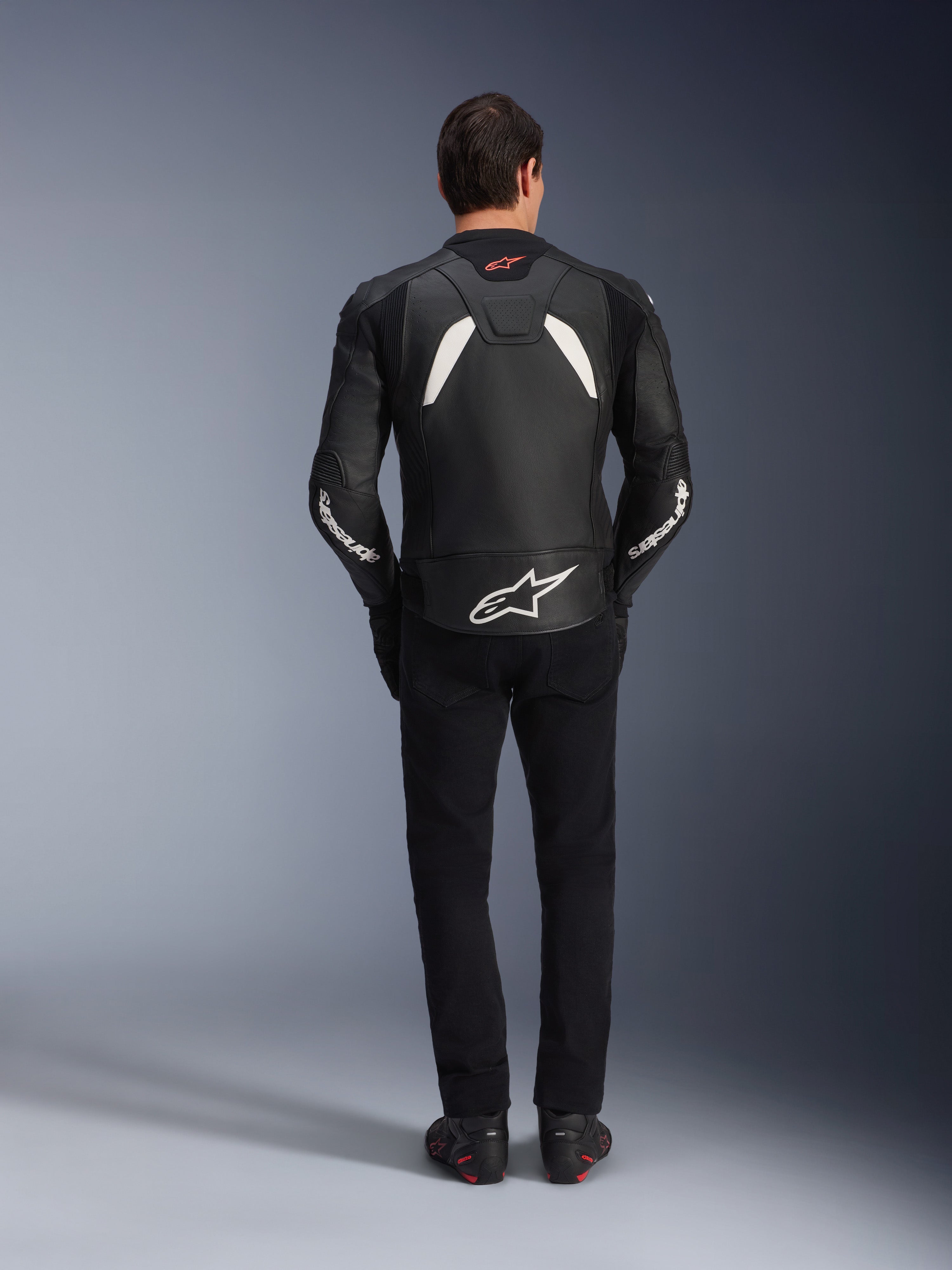 Modèle vu de dos portant le blouson en cuir Alpinestars GP Plus V4 en noir et blanc, associé à un pantalon noir et des bottes de moto, présentant des empiècements blancs sur les épaules et le logo Alpinestars sur les manches et le bas du dos.