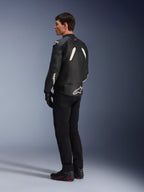 Blouson en cuir Alpinestars GP Plus V4, blouson de sport en cuir, noir et blanc, porté par un mannequin de profil arrière, avec des logos Alpinestars blancs sur le bas du dos et la manche, associé à un pantalon de moto noir et des bottes techniques pour un look de performance sportive.
