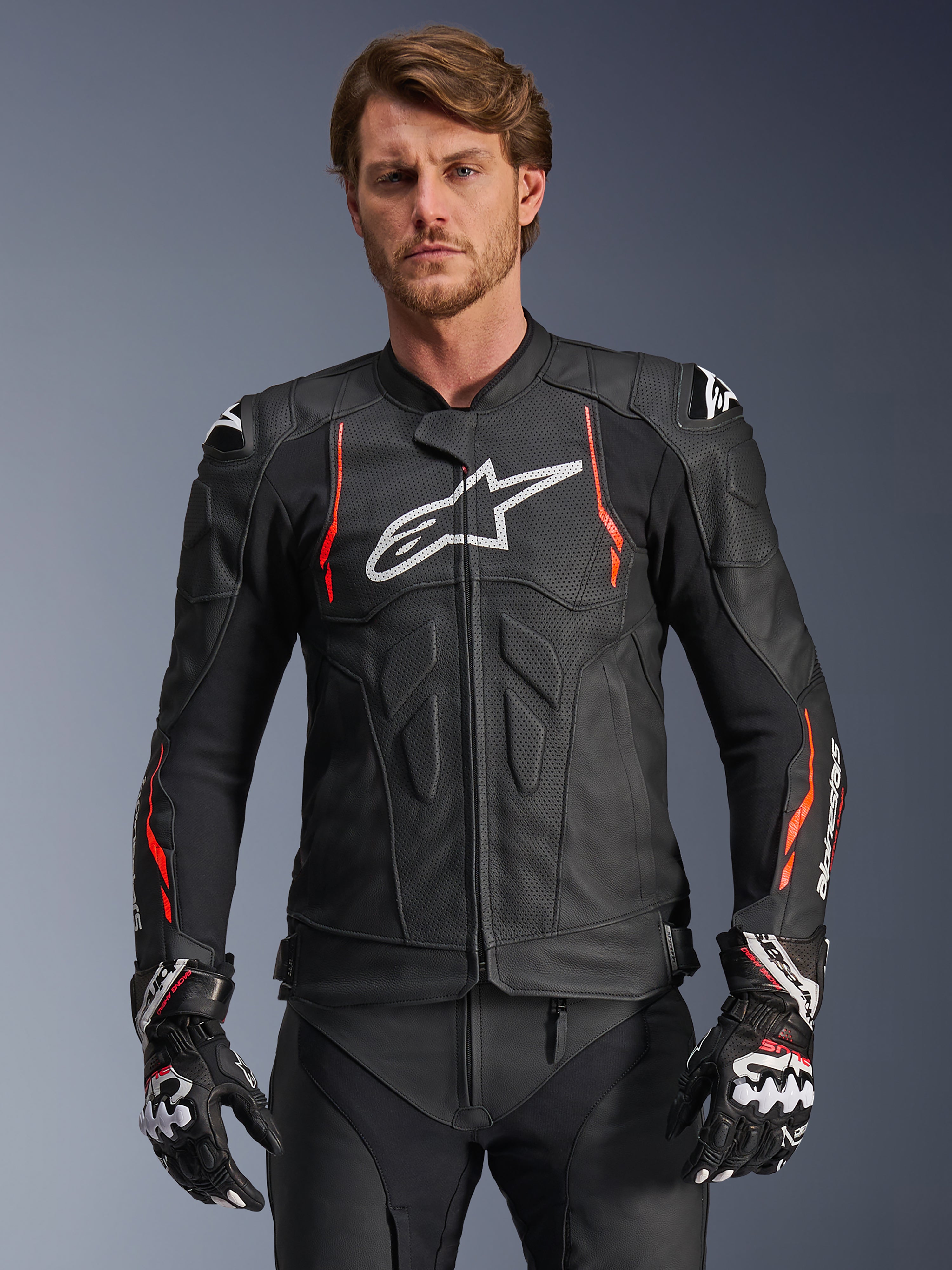 Alpinestars Dusk Airflow leren motorjas, zwart met fluorescerend rode en witte accenten, lederen motorracejas voorzien van uitgebreide perforaties voor ventilatie, wit logo op de borst en externe schoudersliders, gedragen door een model met bijpassende racehandschoenen