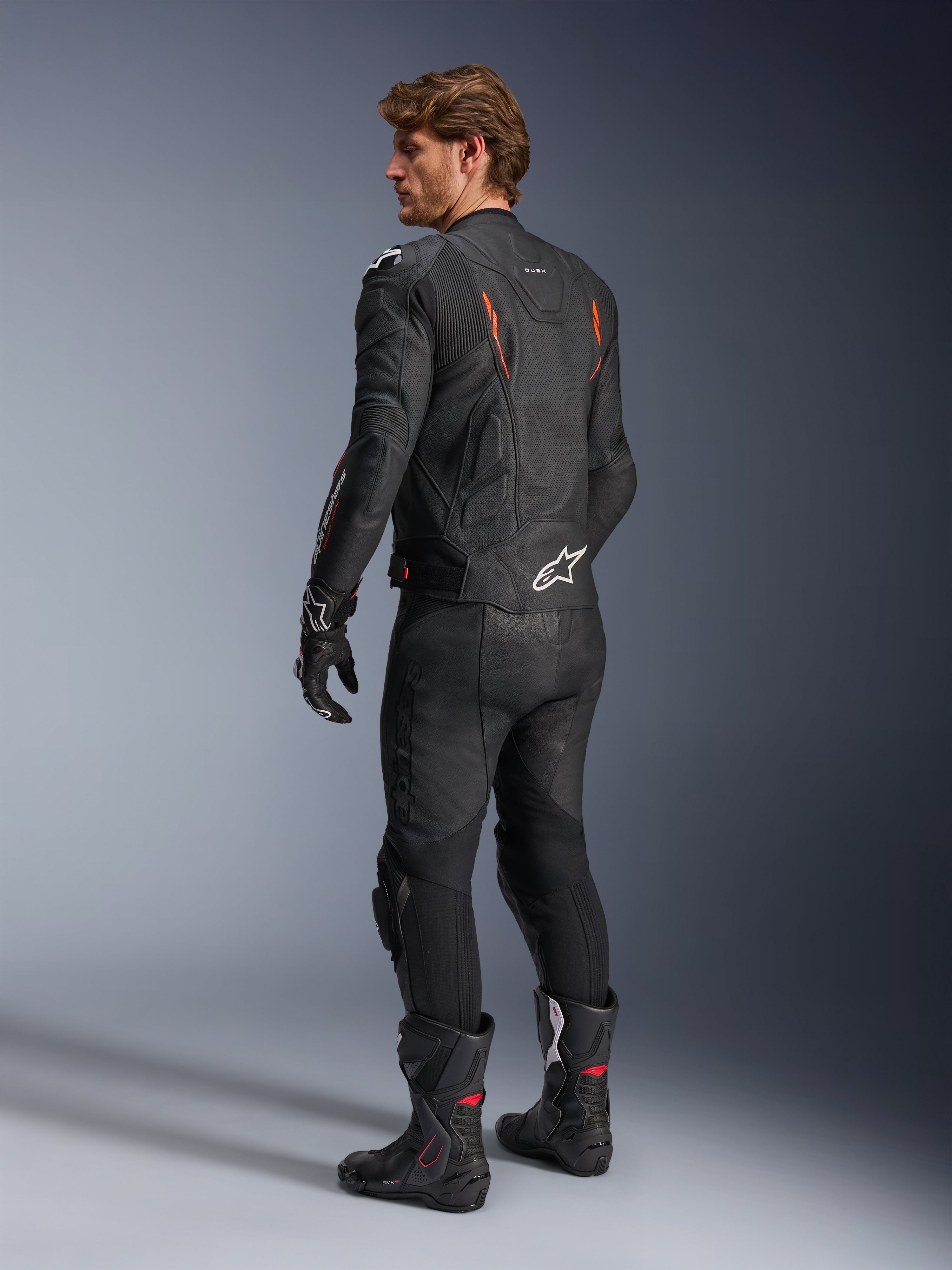Veste en cuir Alpinestars Dusk Airflow, noire avec accents Rouge Fluo, vue arrière d'un pilote portant un équipement de moto complet en cuir avec pantalon, gants et bottes de course assortis, cuir perforé et logos Alpinestars blancs