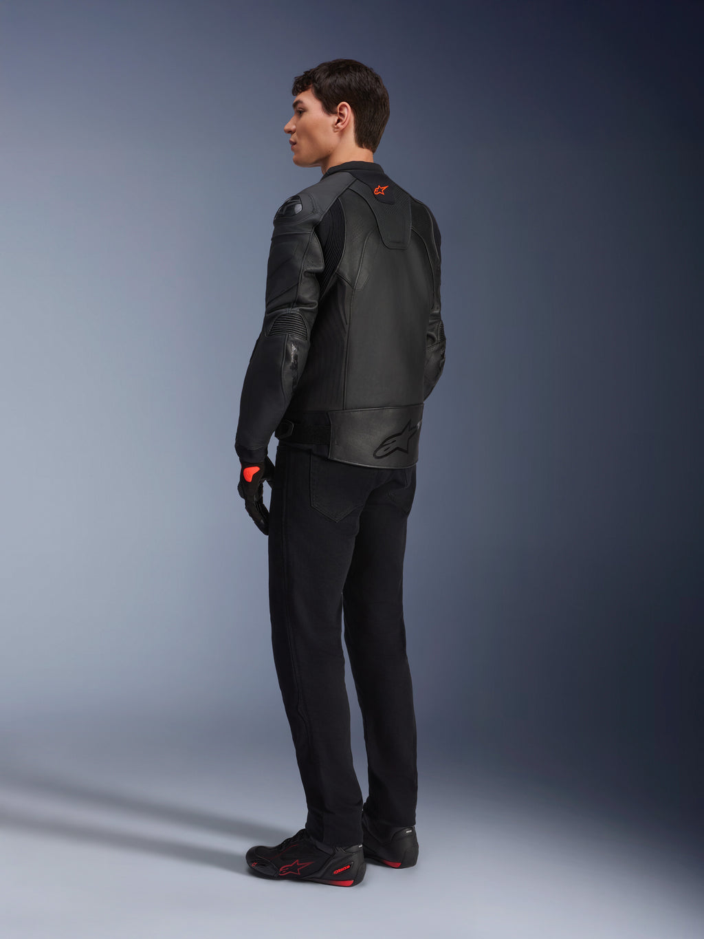 Alpinestars GP Plus R V4 Airflow Lederjacke, Sport-Motorradjacke für Herren, Komplett in Schwarz, Dreiviertel-Rückansicht an einem Model, mit perforierten Lederbahnen, rotem Logo auf dem Rücken und integriertem Schulterschutz, getragen mit schwarzer Hose und Motorradschuhen