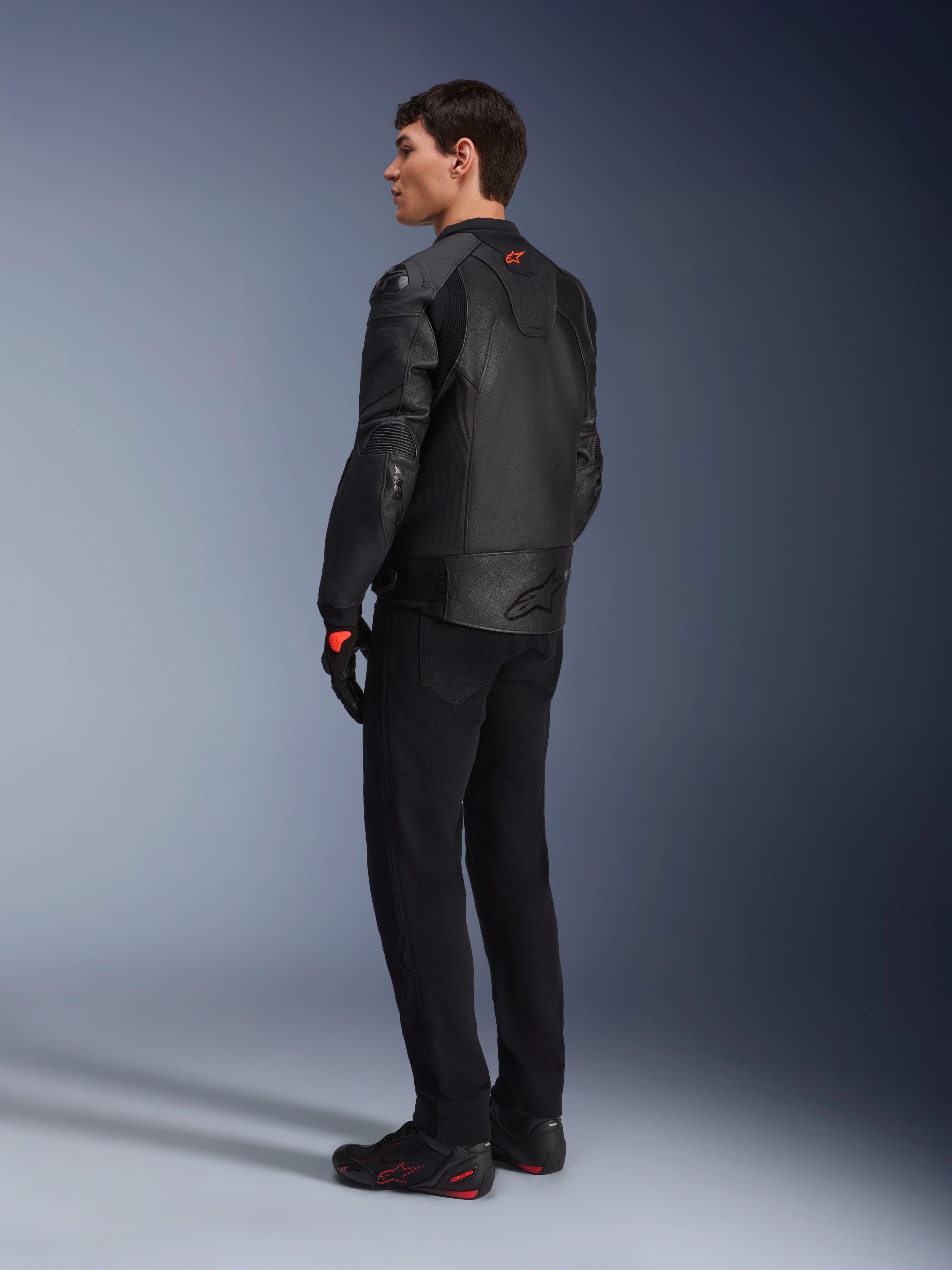 Alpinestars GP Plus R V4 Airflow Lederjacke, Sport-Motorradjacke für Herren, Komplett in Schwarz, Dreiviertel-Rückansicht an einem Model, mit perforierten Lederbahnen, rotem Logo auf dem Rücken und integriertem Schulterschutz, getragen mit schwarzer Hose und Motorradschuhen