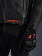 Alpinestars GP Plus R V4 Airflow Lederjacke, Nahaufnahme von schwarzem, perforiertem Leder mit orangefarbenem Grafiktext, kombiniert mit einem schwarz-orangefarbenen Motorradhandschuh mit dem Astars-Logo