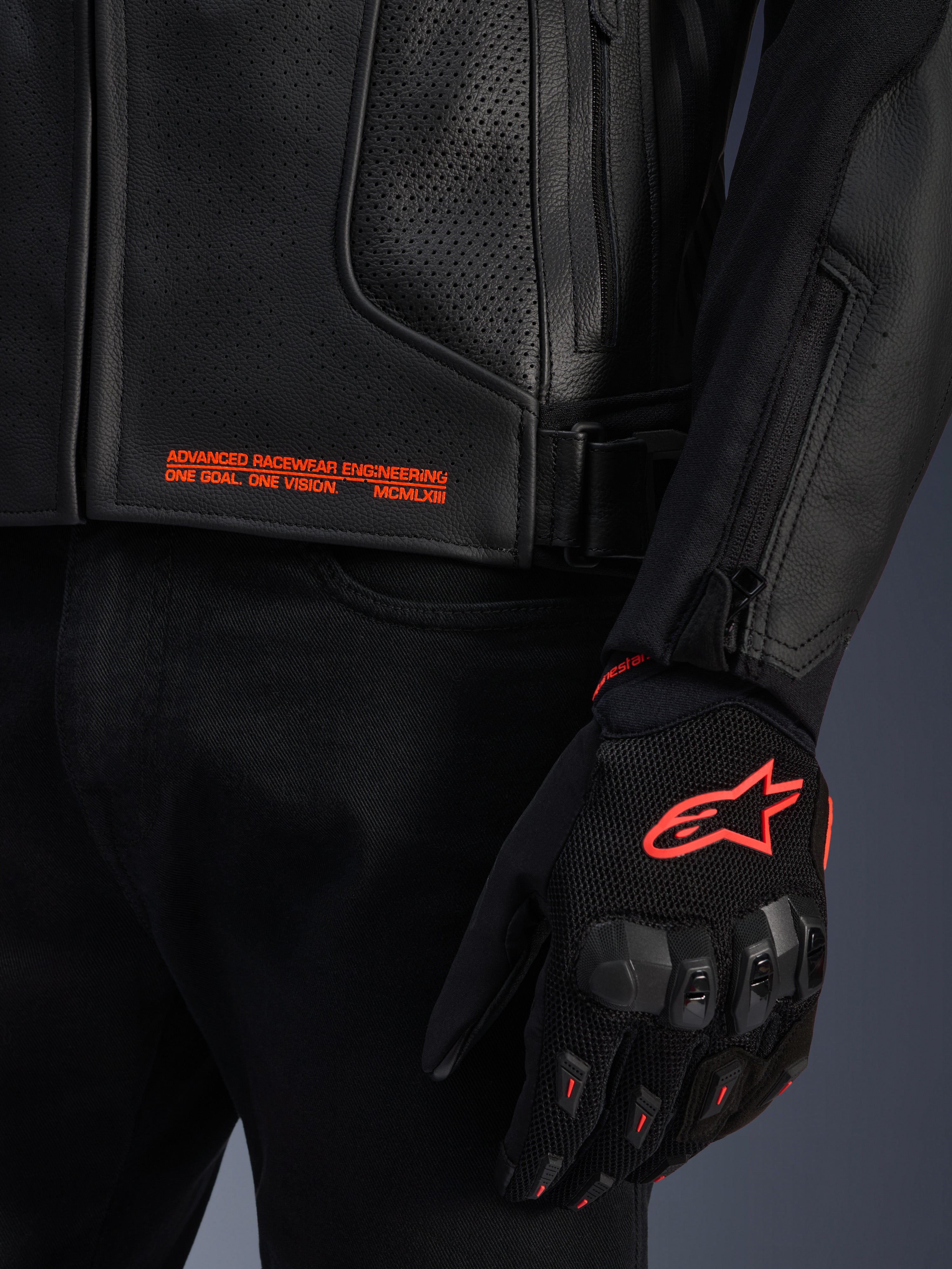 Alpinestars GP Plus R V4 Airflow Lederjacke, Nahaufnahme von schwarzem, perforiertem Leder mit orangefarbenem Grafiktext, kombiniert mit einem schwarz-orangefarbenen Motorradhandschuh mit dem Astars-Logo