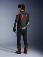 Rückansicht eines Models in der Alpinestars GP Plus R V4 Airflow Lederjacke, Schwarz mit Red Fluo und weißen Akzenten, getragen mit einer schwarzen Hose und schwarzen Motorradstiefeln, mit markanten weißen Logos auf dem Rücken und den Ärmeln, sportliche Lederkonstruktion