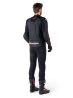 Veste en cuir Alpinestars Faster V3 Airflow, noir et rouge fluo, vue de dos sur un pilote, avec des panneaux en cuir perforé et un marquage distinctif, portée avec un pantalon noir et des bottes de moto, tenue de pilotage sportive.