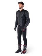 Homme portant la veste en cuir Alpinestars Faster V3 Airflow en Noir Noir, associée à un pantalon noir, des gants de moto noirs et rouges, et des bottes de course noires avec des accents rouges et blancs, équipement de conduite sportive complet