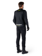 Vue arrière d'un pilote portant la veste en cuir Alpinestars Faster V3 Airflow, noir avec des accents blanc et jaune fluo, associée à un pantalon et des bottes noirs, présentant du cuir perforé pour la ventilation et un marquage 'RACING AHEAD' audacieux sur le bas du dos.