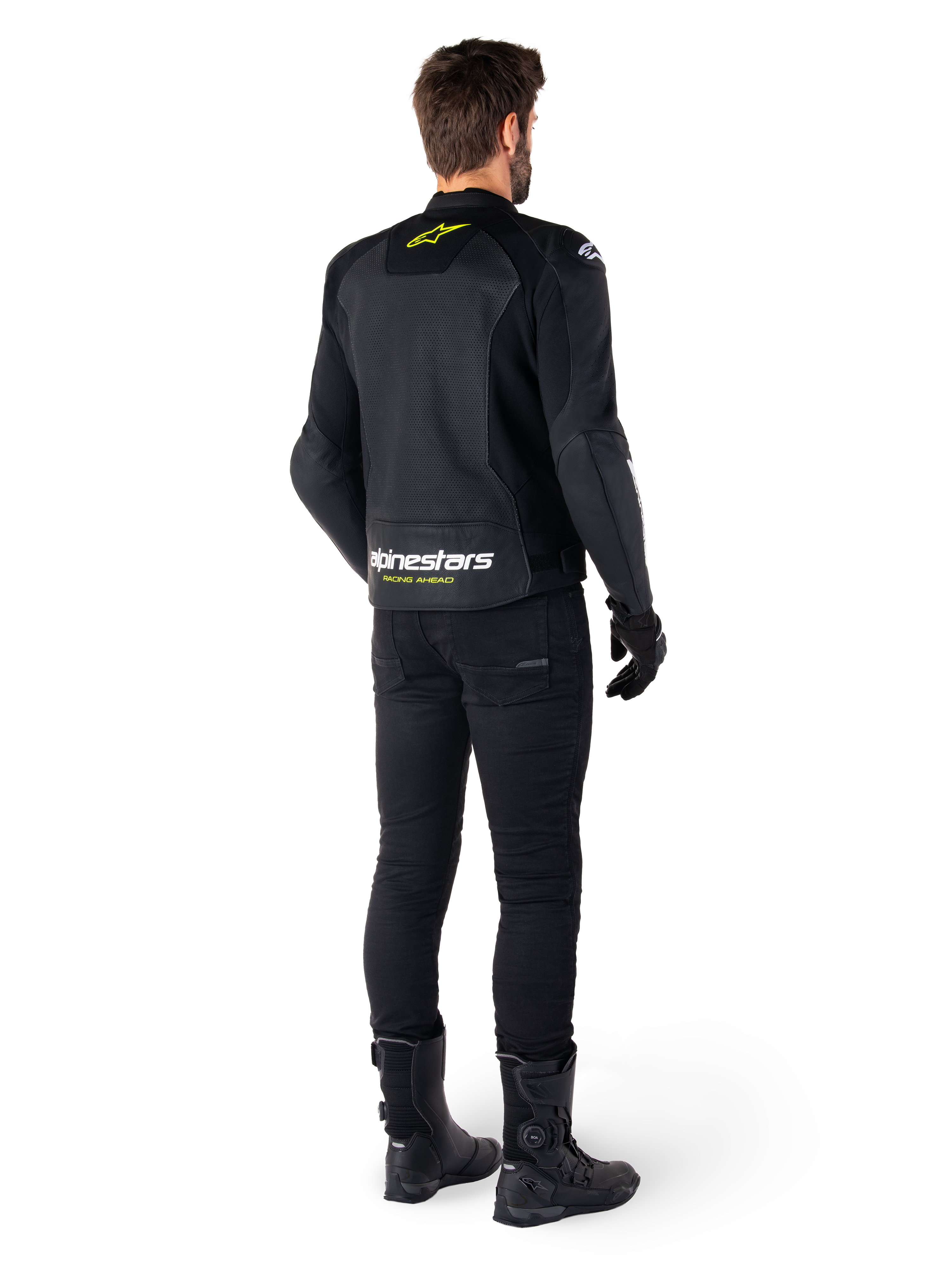 Vue arrière d'un pilote portant la veste en cuir Alpinestars Faster V3 Airflow, noir avec des accents blanc et jaune fluo, associée à un pantalon et des bottes noirs, présentant du cuir perforé pour la ventilation et un marquage 'RACING AHEAD' audacieux sur le bas du dos.