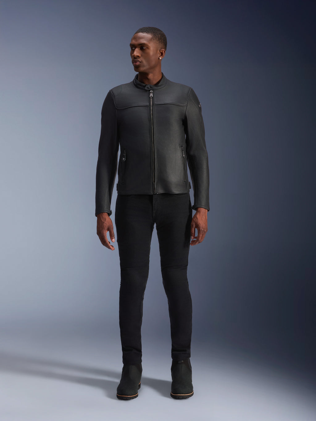 Modèle masculin portant la veste en cuir Alpinestars Blacktrack, cuir de bovin noir de première qualité, design de motard minimaliste moderne avec une coupe sur mesure, associé à un pantalon de conduite et des bottes noirs, style urbain contemporain