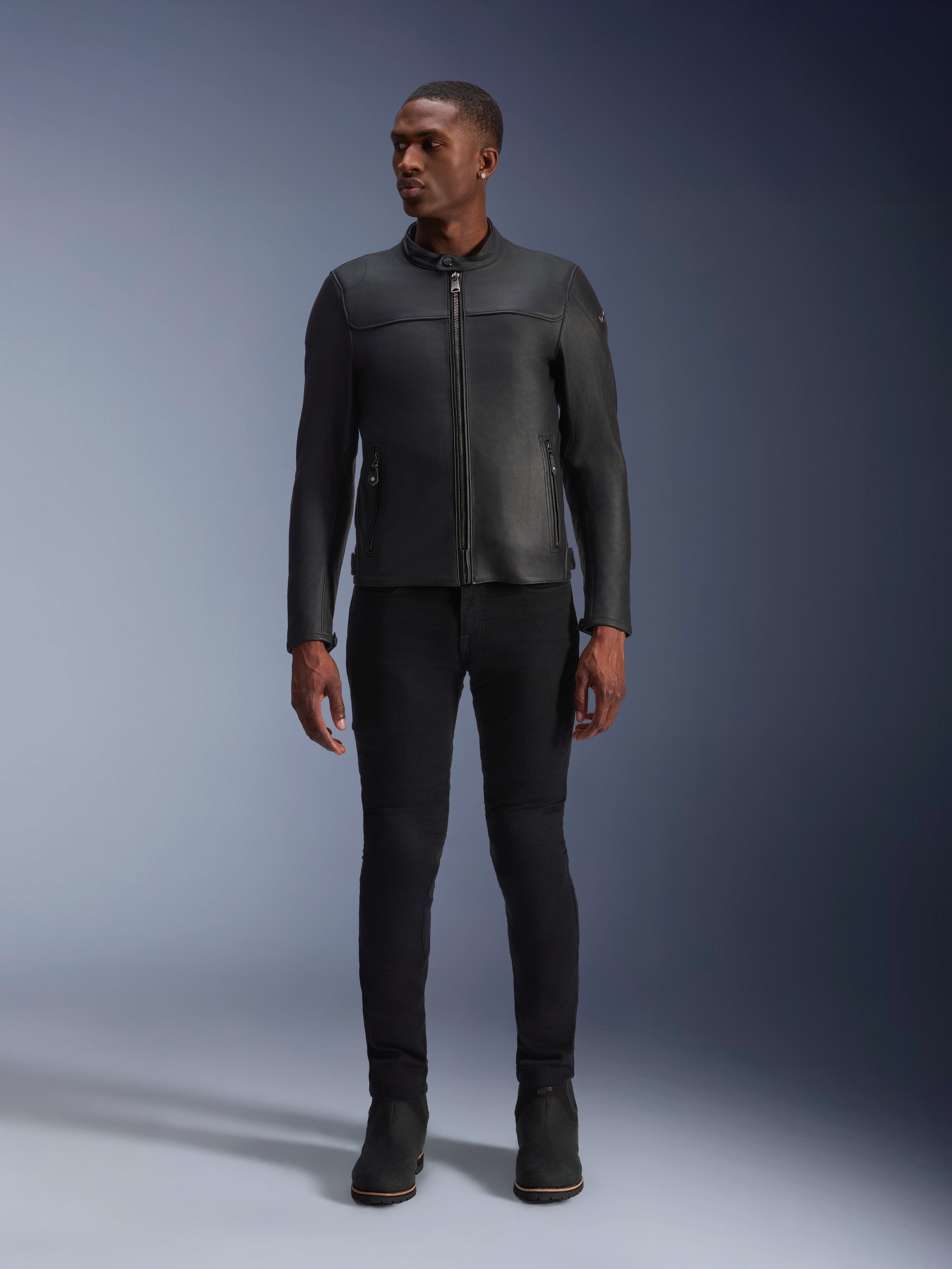Modèle masculin portant la veste en cuir Alpinestars Blacktrack, cuir de bovin noir de première qualité, design de motard minimaliste moderne avec une coupe sur mesure, associé à un pantalon de conduite et des bottes noirs, style urbain contemporain