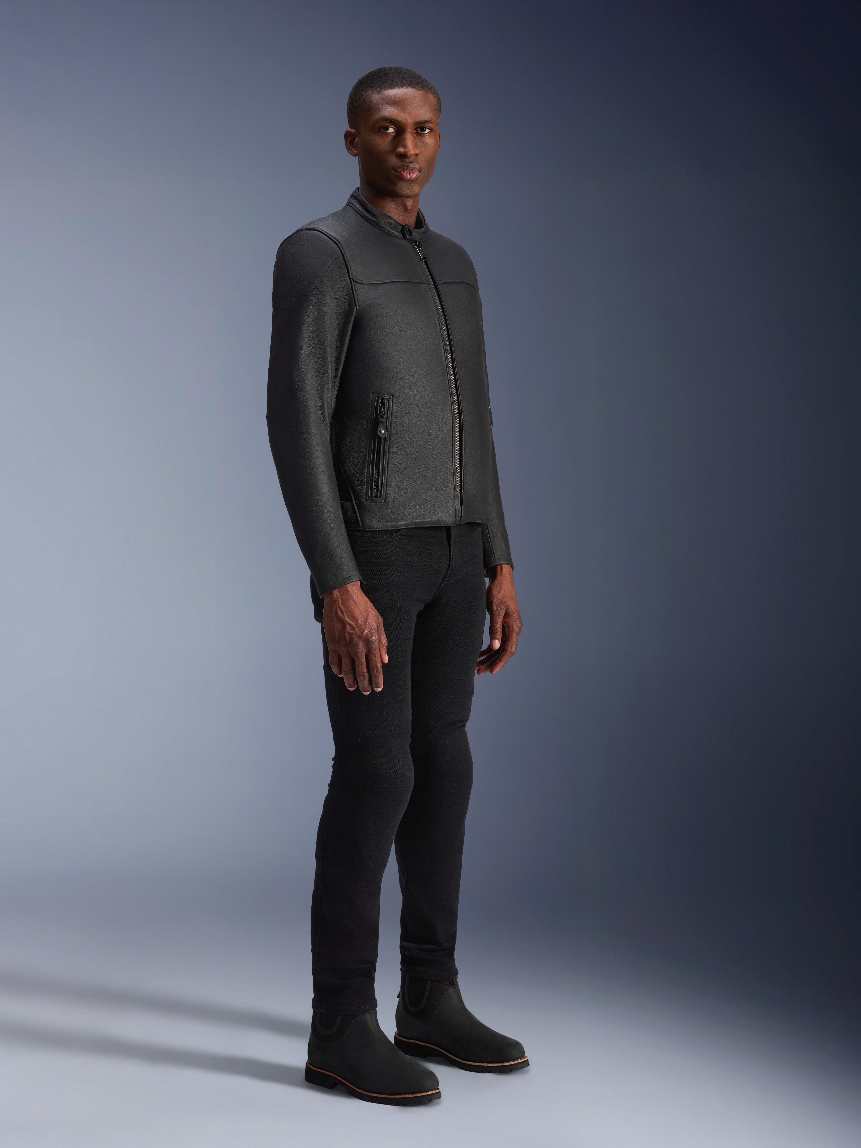 Modèle masculin portant la veste en cuir Alpinestars Blacktrack, noir sur noir, style motard moderne avec cuir premium et détails de zips métalliques, associé à un pantalon et des bottes noirs pour un look urbain contemporain