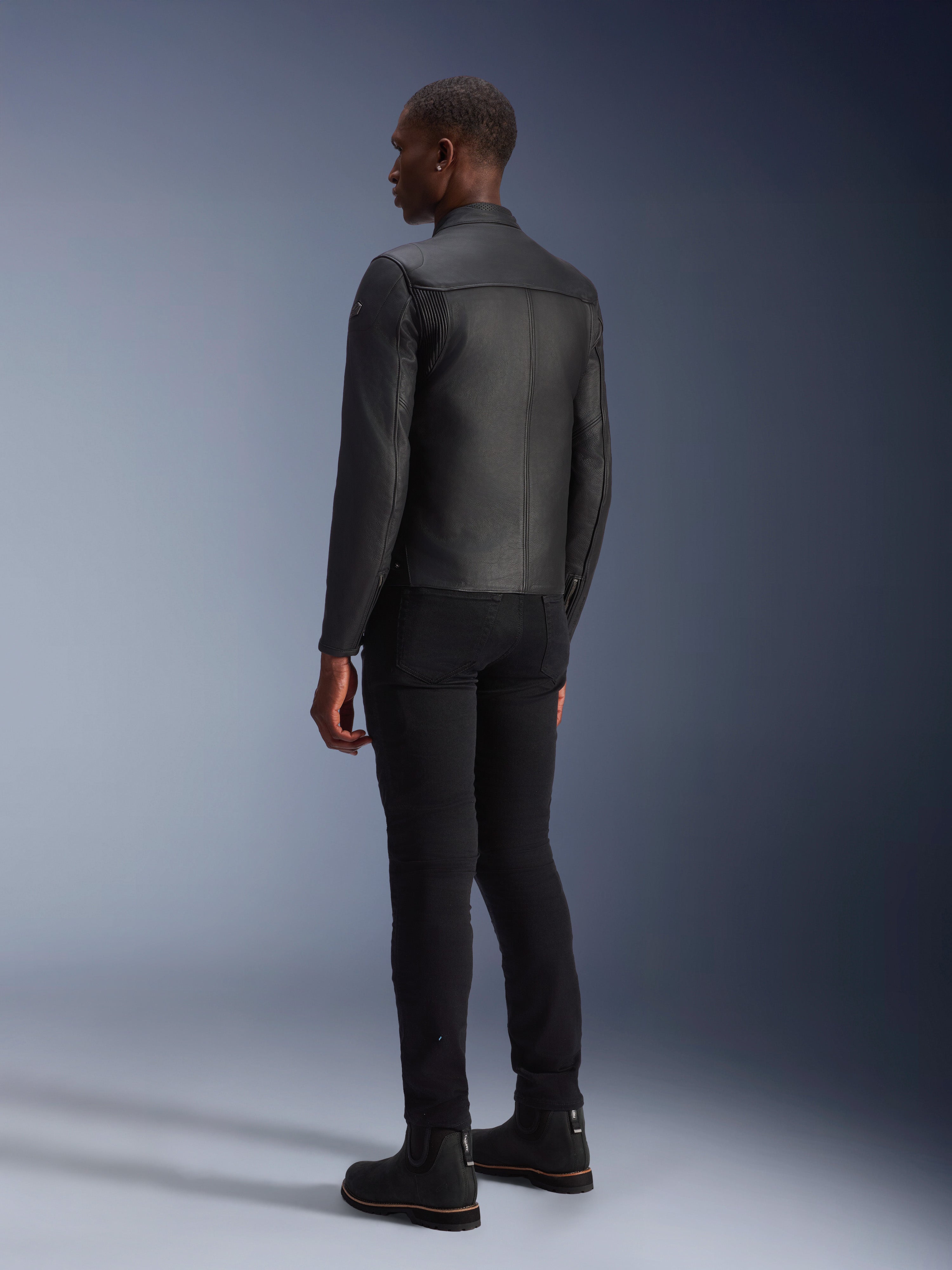 Veste en cuir Alpinestars Blacktrack, noir, vue de dos montrant la coupe ajustée et les panneaux en cuir accordéon sur les omoplates, portée avec un pantalon noir et des bottes noires, veste de moto en cuir de bovin haut de gamme