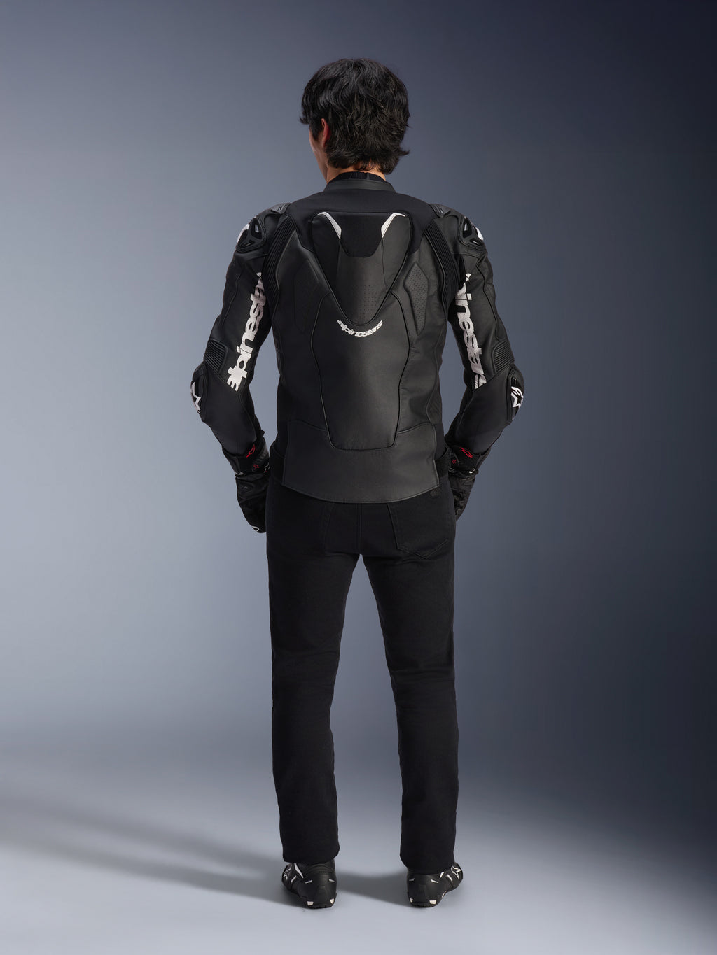 Alpinestars Atem V5 Leren Jas, motorracejas, zwart en wit, achteraanzicht op rijder, voorzien van aerodynamische snelheidsbult en witte Alpinestars-branding op mouwen en rug, technische lederen constructie, gedragen met zwarte broek en motorlaarzen