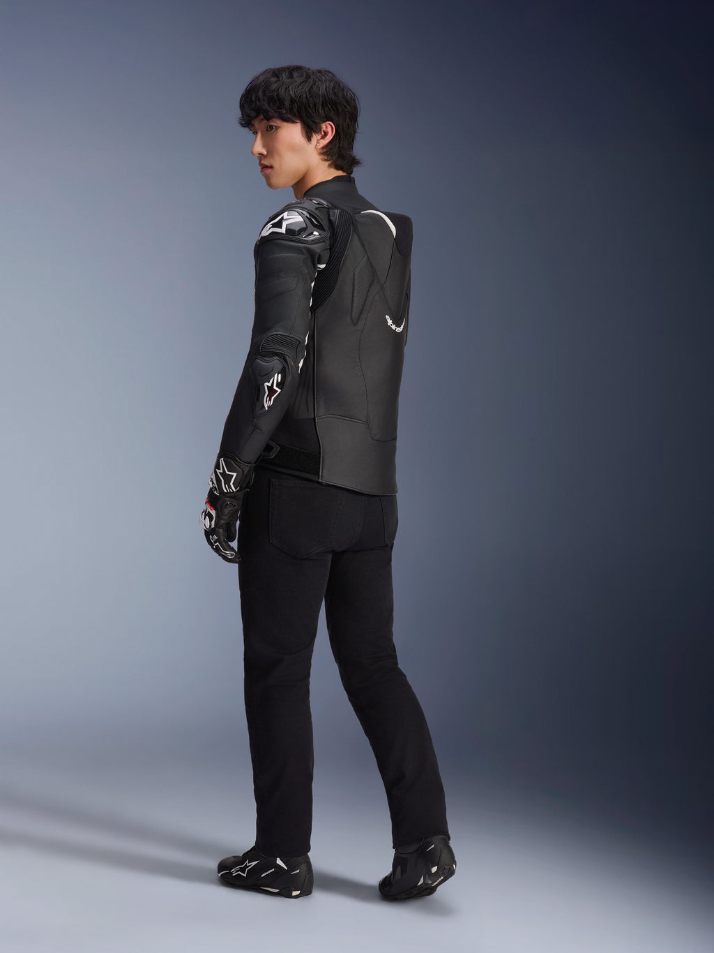 Mannelijk model draagt de Alpinestars Atem V5 leren jas in zwart en wit, driekwart achteraanzicht met de aerodynamische snelheidsbult en DFS RR-protectoren, gecombineerd met een zwarte broek, racehandschoenen en laarzen, professionele motorracekleding.
