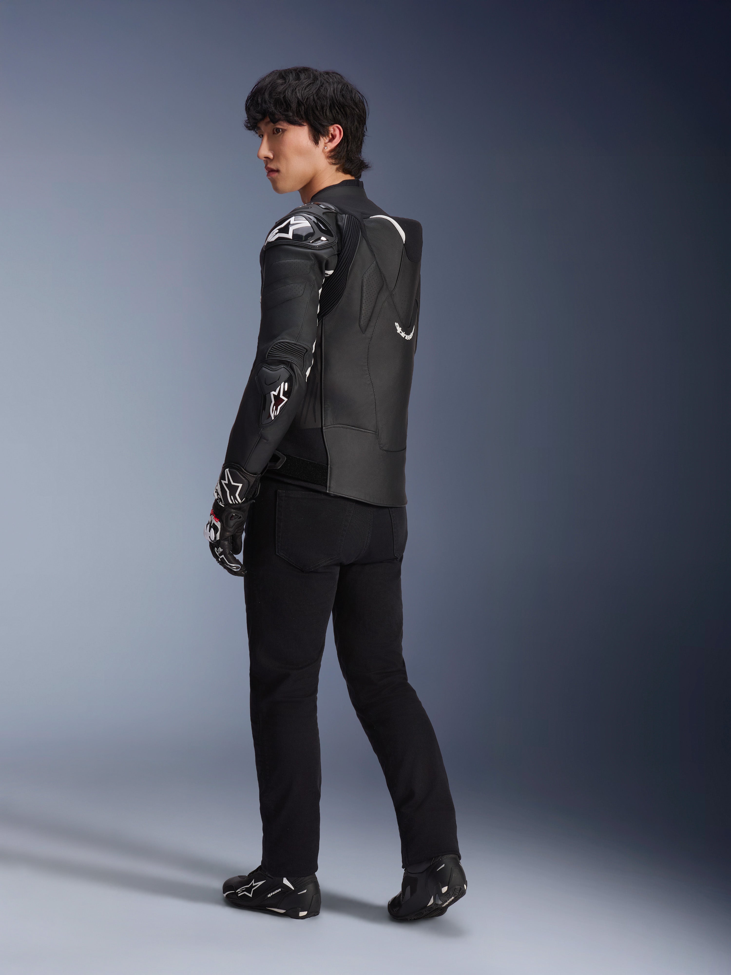 Mannelijk model draagt de Alpinestars Atem V5 leren jas in zwart en wit, driekwart achteraanzicht met de aerodynamische snelheidsbult en DFS RR-protectoren, gecombineerd met een zwarte broek, racehandschoenen en laarzen, professionele motorracekleding.