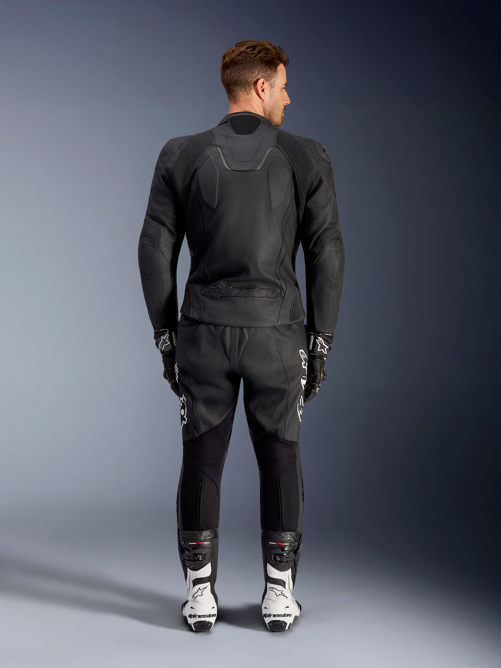 Achteraanzicht van een man die de Alpinestars Caliber Leren Jas in het zwart draagt, voorzien van een reliëflogo en geperforeerde lederen panelen, gecombineerd met een zwarte lederen motorbroek, zwarte handschoenen en witte racelaarzen.