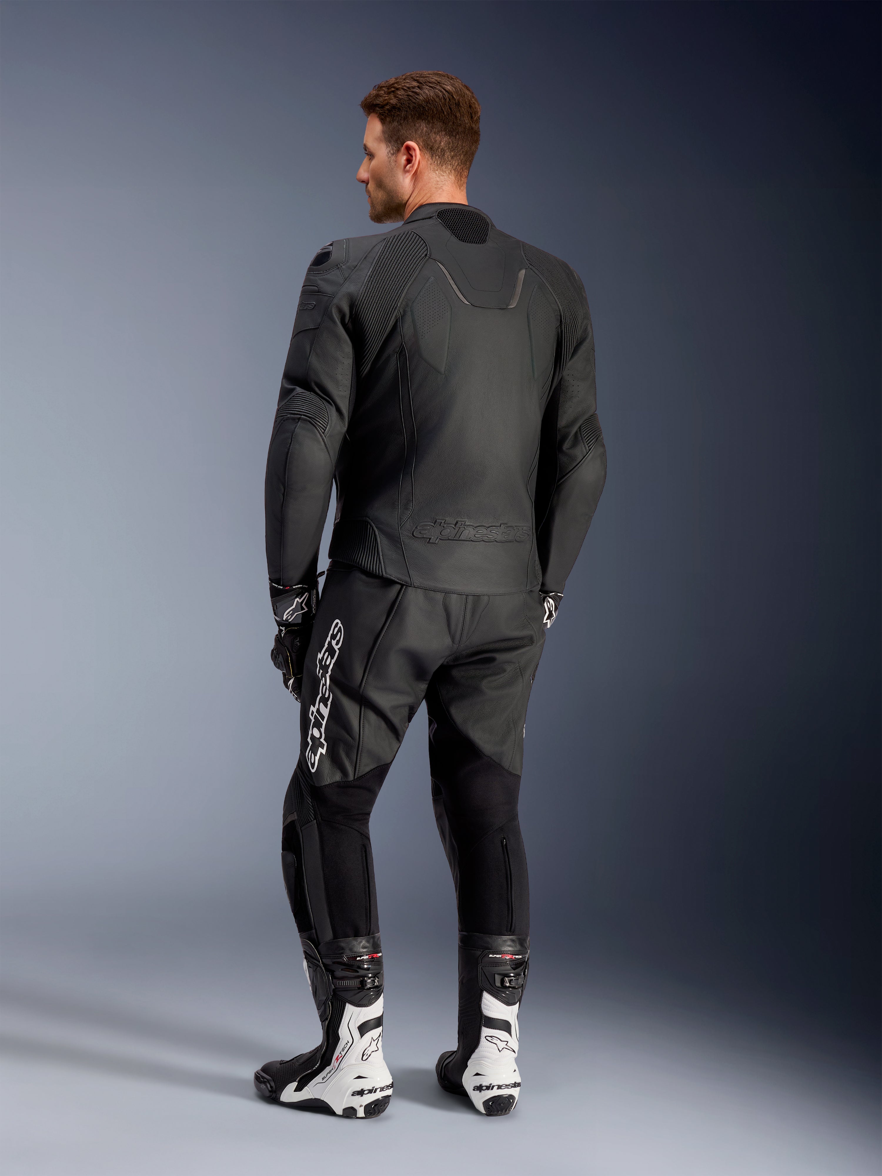 Alpinestars Caliber leren jas, zwarte motorjas, achteraanzicht, voorzien van geperforeerd leer en accordeon stretch-inzetten in de taille en op de schouders, logo in reliëf, gedragen met zwarte leren broek en witte racelaarzen