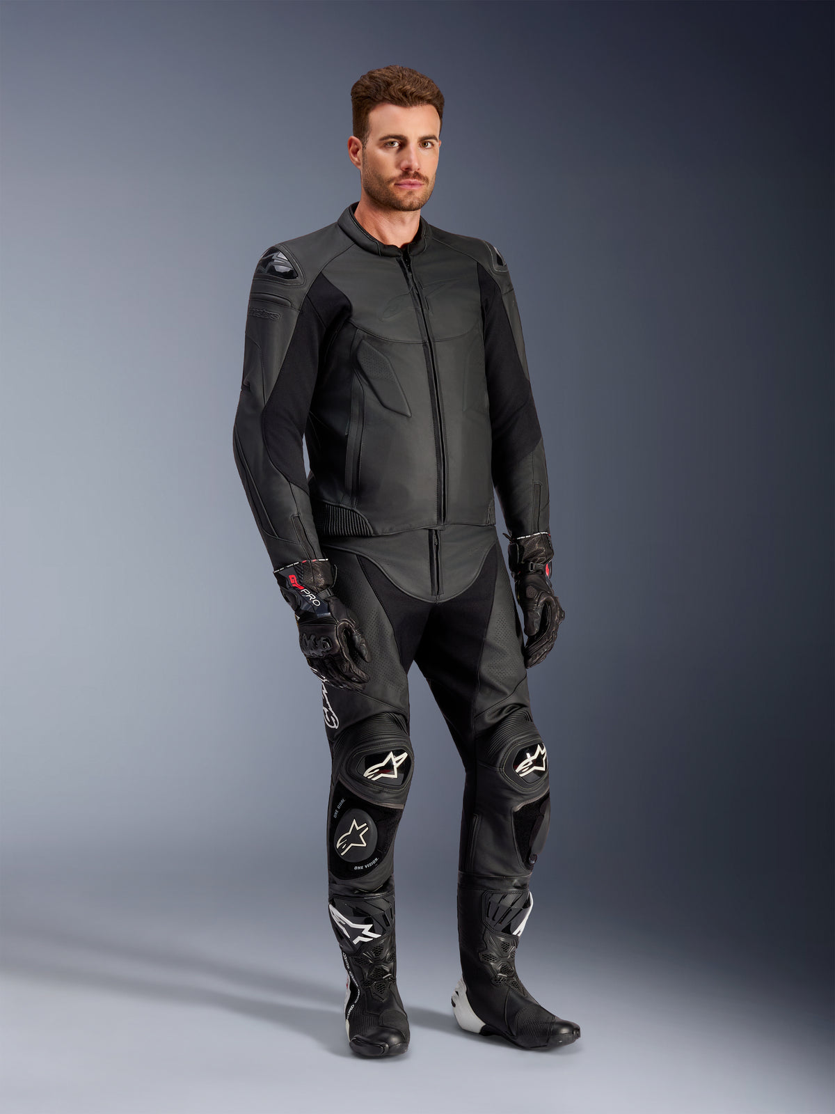 Modèle masculin portant la veste en cuir Alpinestars Caliber noire, associée à un pantalon de moto en cuir noir assorti, des gants de course et des bottes, présentant une tenue complète d'équipement de route sportif.