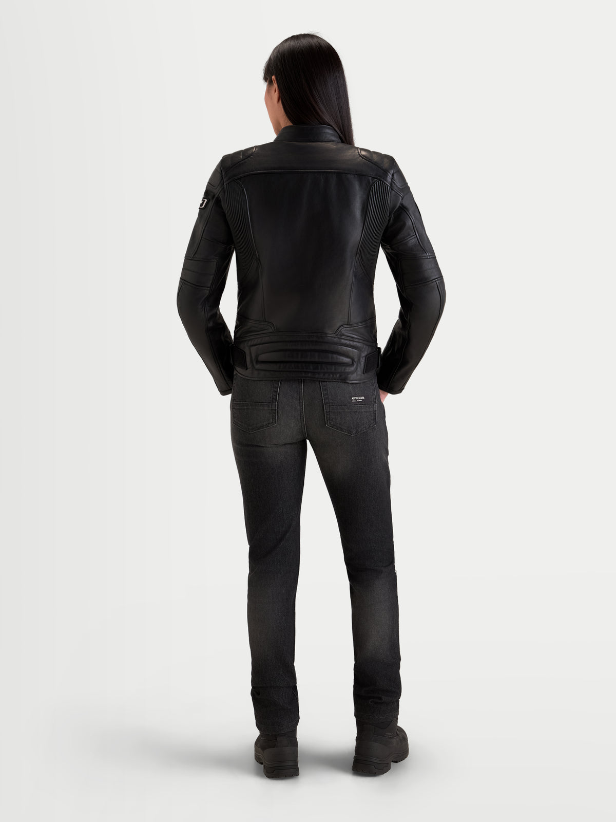 Alpinestars Revea Veste en cuir, blouson de moto urbain en cuir noir pour femme, vue de dos sur un modèle portant un jean foncé et des bottes, construction en cuir de bovin pleine fleur