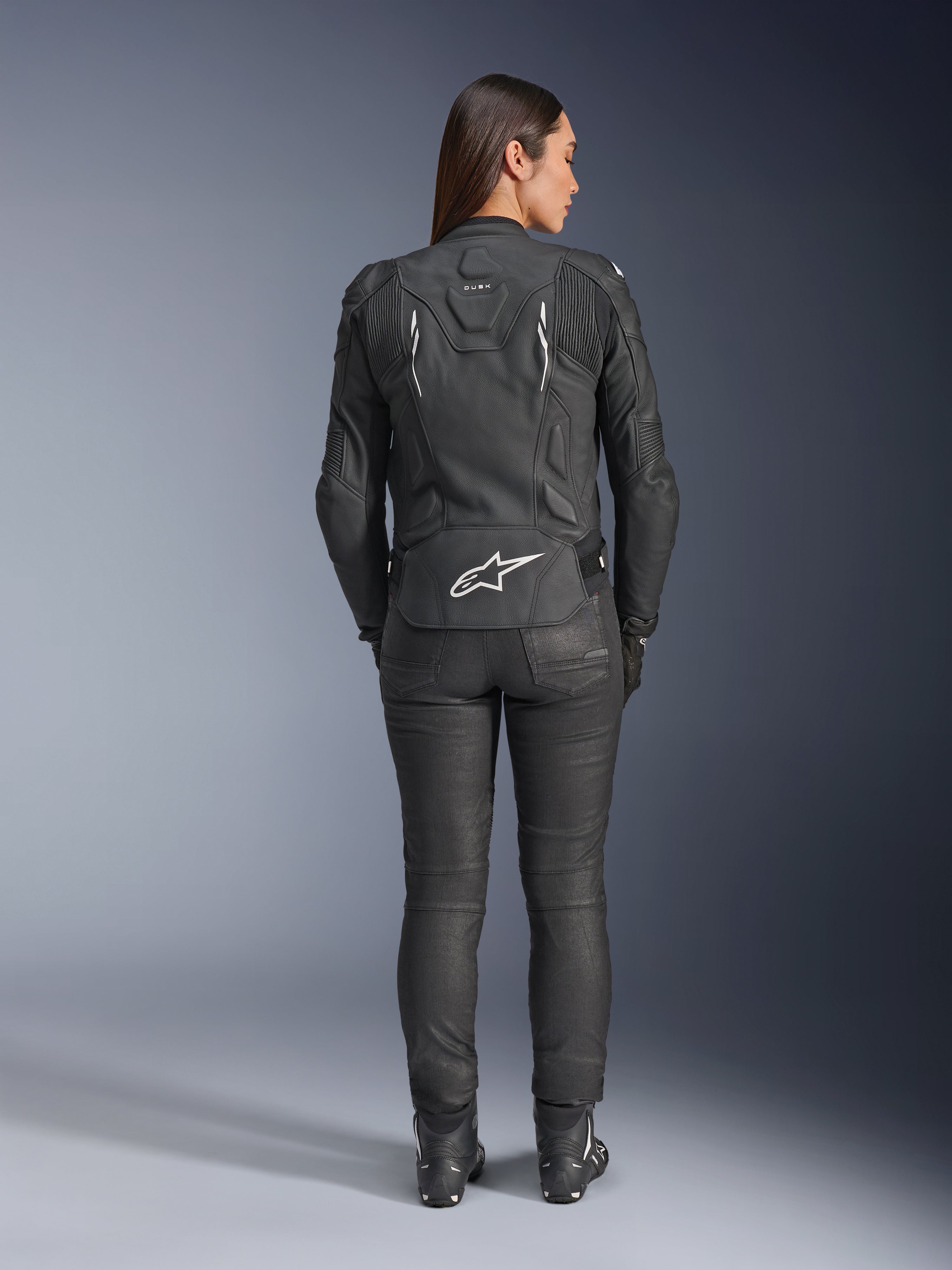 Alpinestars Stella Dusk leren jas voor dames, zwart, achteraanzicht met wit Alpinestars-logo op de onderrug en 'DUSK'-branding op de bovenrug, gedragen met zwarte motorbroek en laarzen, toont de aansluitende damespasvorm en beschermende leren constructie.