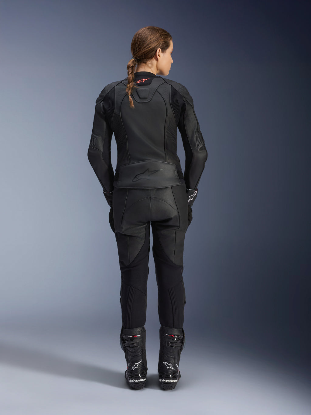 Alpinestars Stella GP Plus V4 leren damesjas, volledig zwart, volledig achteraanzicht van een vrouwelijke motorrijder met bijpassende leren broek en laarzen, strak minimalistisch ontwerp met een klein rood logo op de nek, sportief getailleerd silhouet voor dames