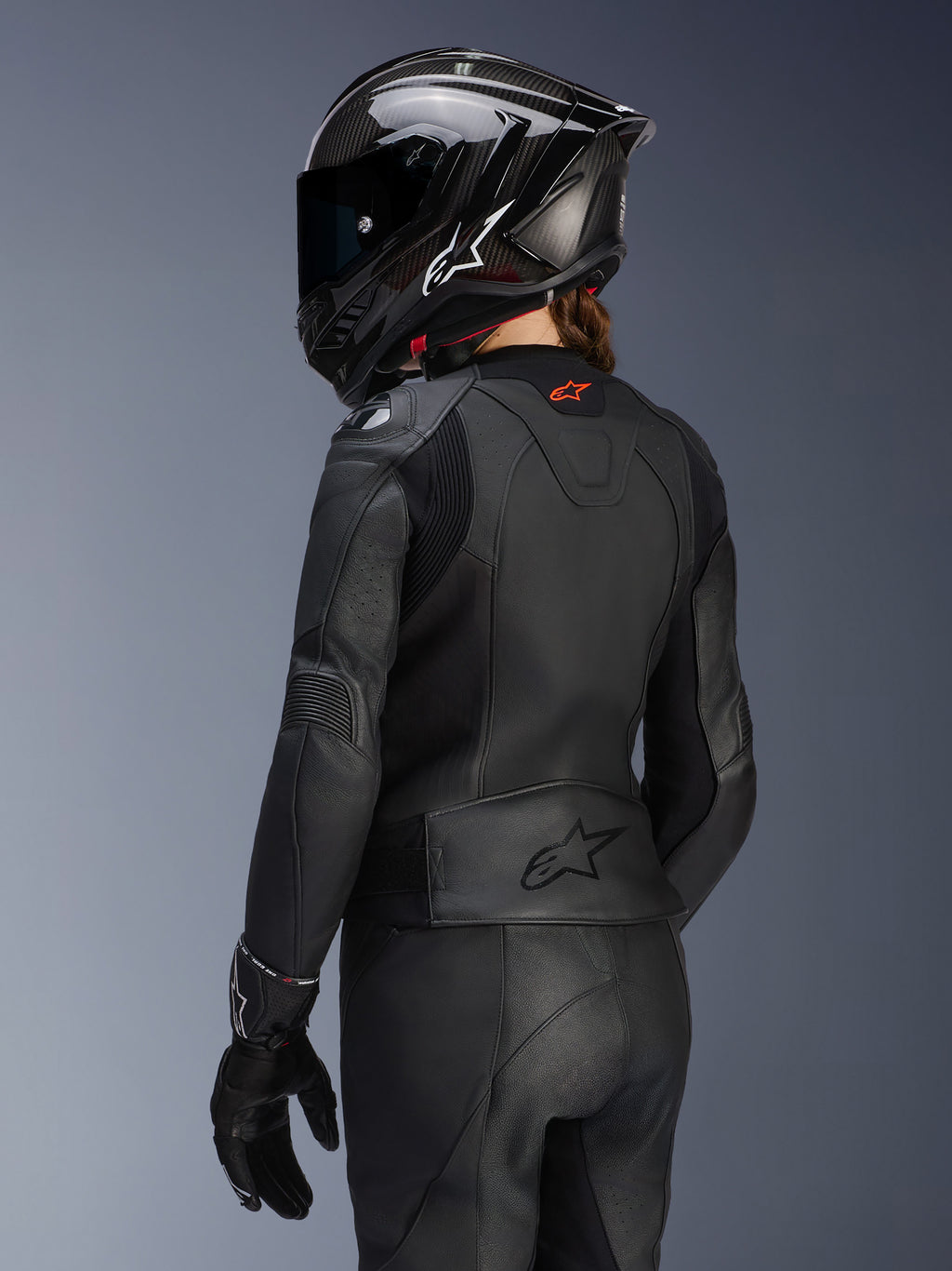 Vrouwelijke motorrijder van achteren, Alpinestars Stella GP Plus V4 Leren Damesjas in het zwart, bijpassende leren broek en handschoenen, zwarte carbon-stijl integraalhelm, aerodynamische pasvorm en reliëflogo op de onderrug