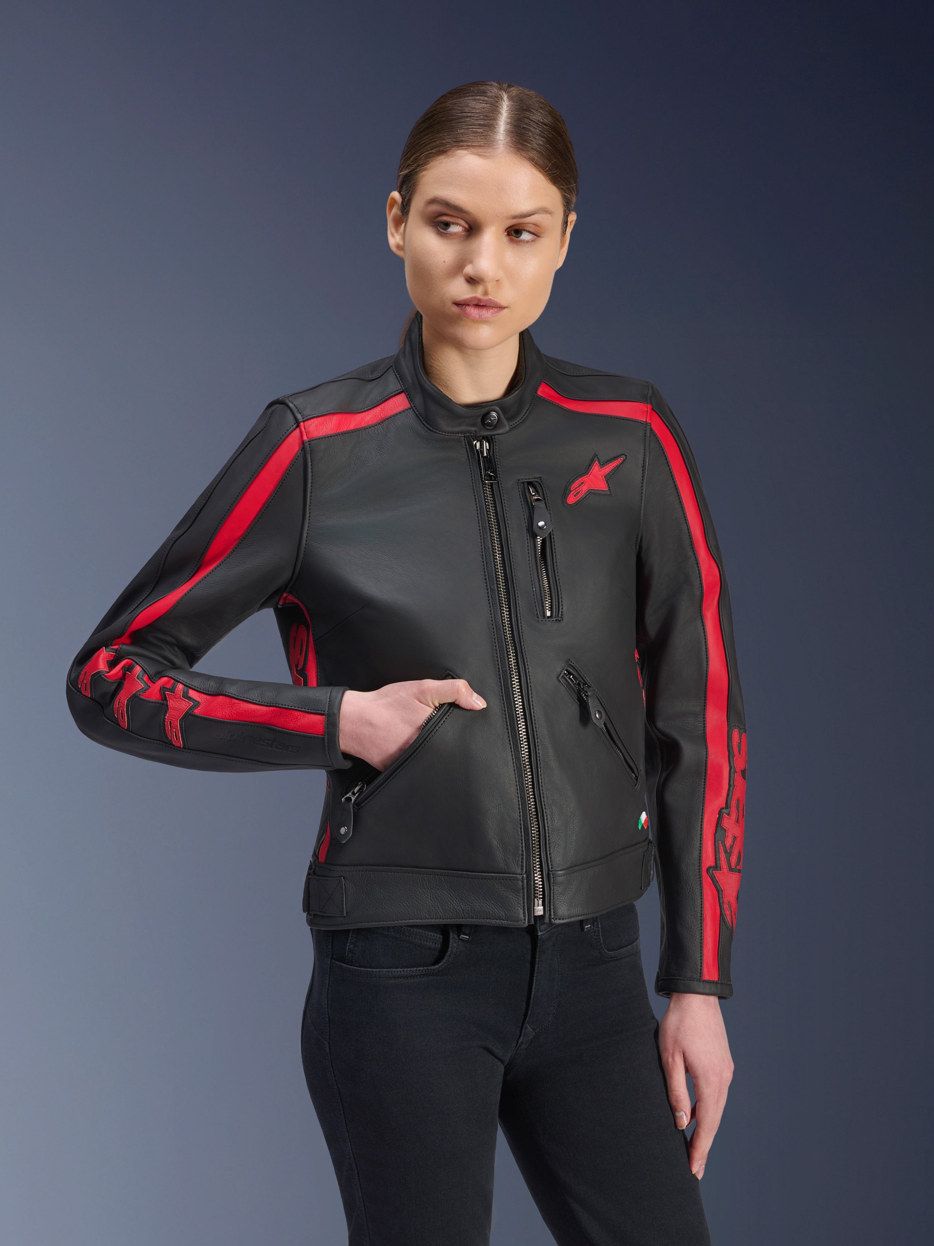 Alpinestars Stella Dyno leren jas, dames motorjas, zwart met rood, gedragen door een model met zwarte jeans, rode racing strepen op de schouders en mouwen met opvallende branding, hoogwaardige leren constructie, op erfgoed geïnspireerd ontwerp voor stedelijke ritten