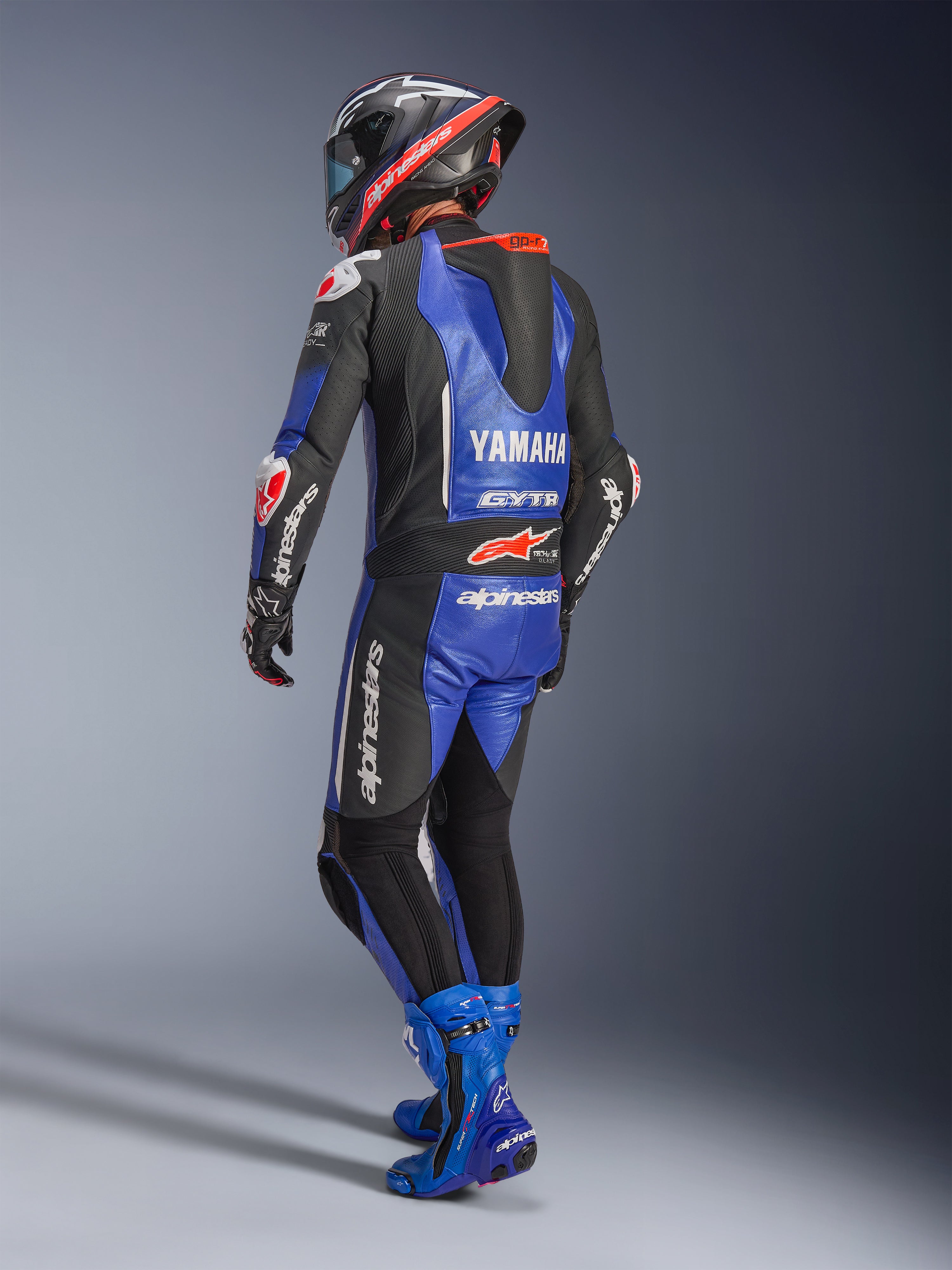 Alpinestars Yamaha GP-R7 Einteiliger Lederanzug, Metallic-Blau, Schwarz und Weiß, Rückansicht mit aerodynamischem Rennhöcker und Yamaha-Branding, getragen mit passenden blauen Rennstiefeln und Helm, professioneller Performance-Lederanzug für den Rennsport