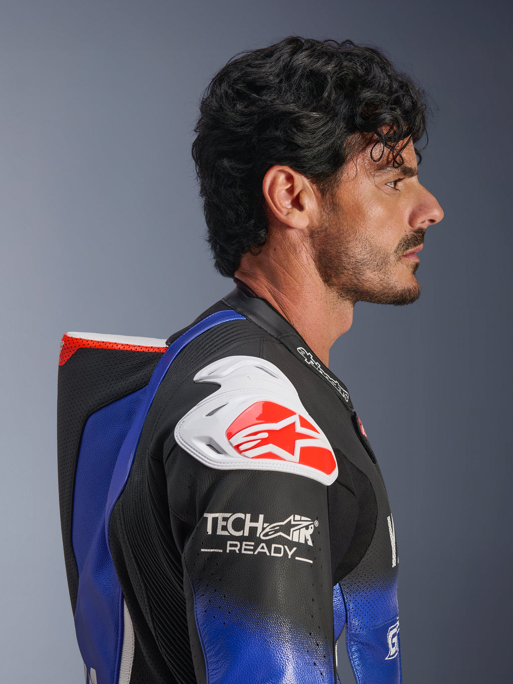 Alpinestars Yamaha GP-R7 einteiliger Lederanzug, Metallic-Blau und Schwarz mit weißen und roten Akzenten, Seitenprofil zeigt den aerodynamischen Rückenhöcker und den weißen Schulterprotektor mit rotem Alpinestars-Logo, Tech-Air Ready-Branding am Ärmel, hochwertiges perforiertes Rennleder.