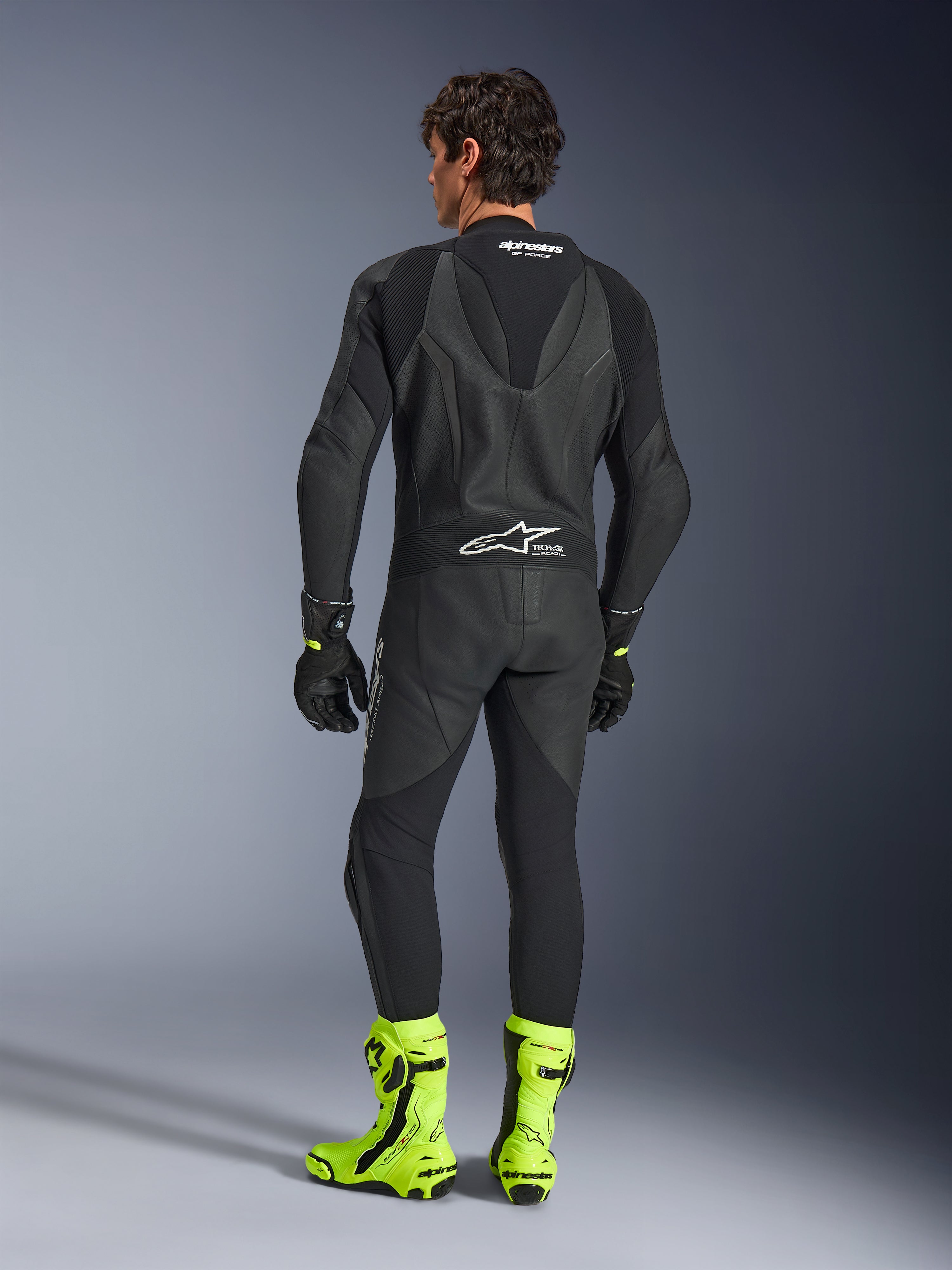 Combinaison en cuir Alpinestars GP Force V2 - 1 pièce, noir, vue de dos portée par un mannequin, avec bosse aérodynamique et marquage Tech-Air Ready, assortie de gants noirs et de bottes de course jaune fluorescent vif, équipement de piste professionnel