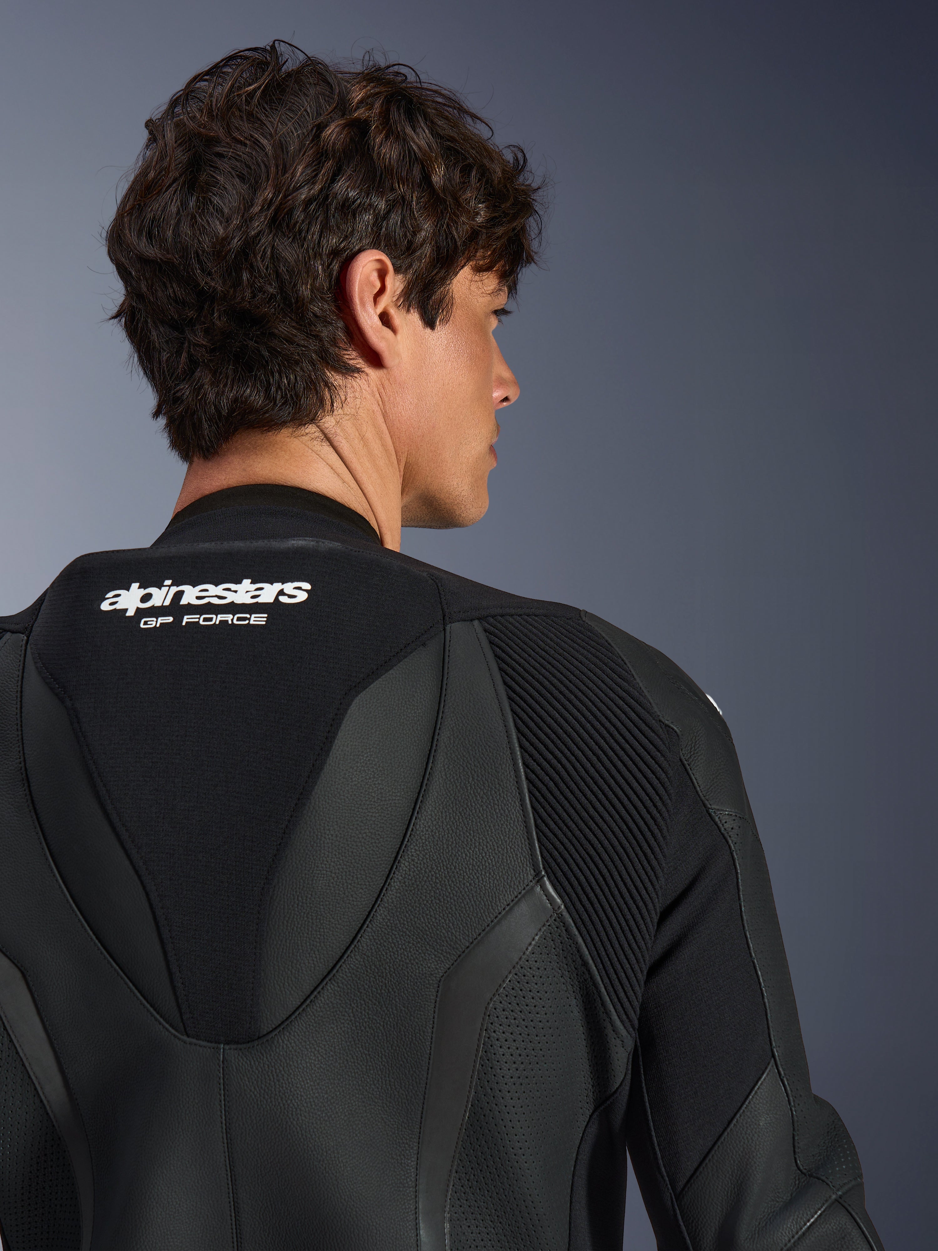 Alpinestars GP Force V2 Leather Suit 1 pièce, noir, vue rapprochée du haut du dos et de la bosse aérodynamique, avec logos blancs, sections en cuir perforé et panneaux extensibles en accordéon pour le pilotage sportif.