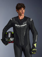 Combinaison en cuir Alpinestars GP Force V2, combinaison de course de moto une pièce noire avec panneaux en cuir perforé et logo blanc, portée par un mannequin tenant un casque et des gants assortis, éclairage studio