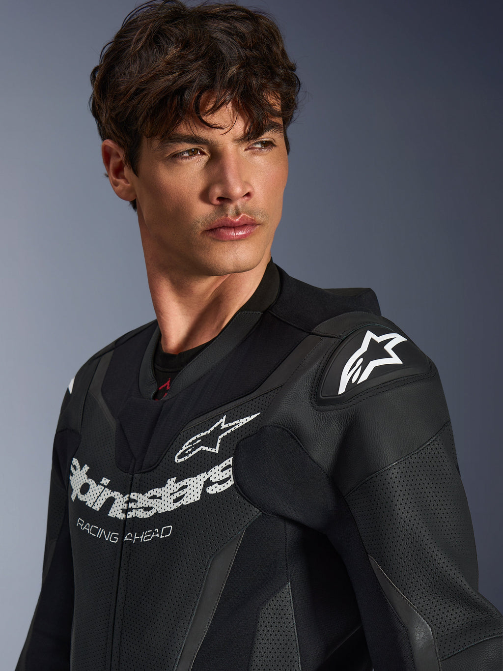 Alpinestars GP Force V2 Leather Suit, gros plan d'un homme portant la combinaison noire, construction en cuir perforé, logo Alpinestars avec marquage « RACING AHEAD » sur la poitrine, logo étoile blanche sur les sliders d'épaules