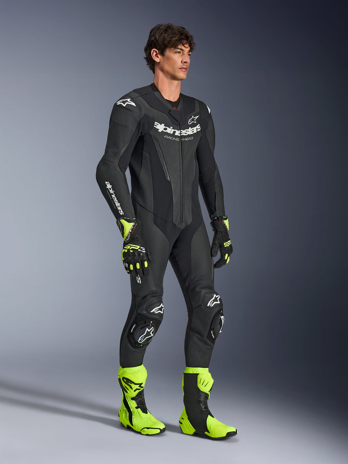 Homme portant la combinaison une pièce en cuir Alpinestars GP Force V2 en noir, avec logos Alpinestars blancs, sections en cuir perforé et protections intégrées, associée à des bottes et des gants de course jaune fluo et noirs