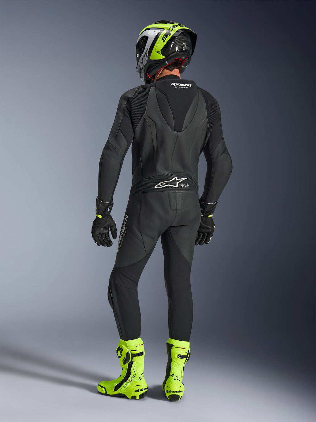 Vue arrière d'un pilote portant la combinaison en cuir Alpinestars GP Force V2 une pièce en noir, montrant la bosse aérodynamique et le marquage Tech-Air Ready, associée à un casque noir et jaune fluo et des bottes de course jaune vif.