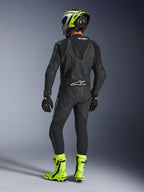 Vue arrière d'un pilote portant la combinaison en cuir Alpinestars GP Force V2 une pièce en noir, montrant la bosse aérodynamique et le marquage Tech-Air Ready, associée à un casque noir et jaune fluo et des bottes de course jaune vif.