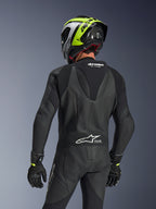 Combinaison en cuir 1 pièce Alpinestars GP Force V2, noire, vue arrière montrant la bosse aérodynamique et la structure en cuir perforé, portée avec un casque de course noir et jaune fluo et des gants noirs, avec panneaux extensibles en accordéon et logo blanc Tech-Air Ready.