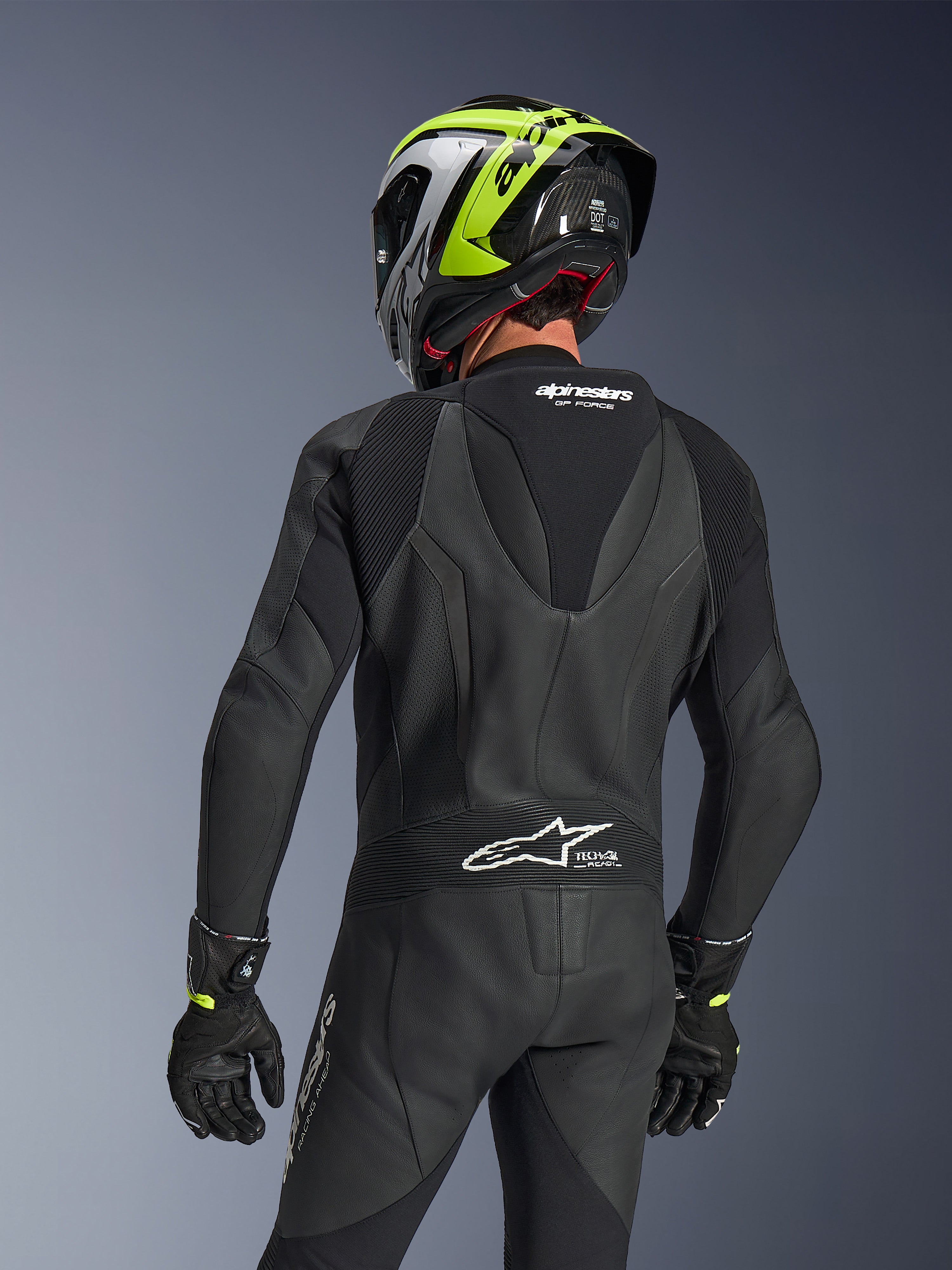 Combinaison en cuir 1 pièce Alpinestars GP Force V2, noire, vue arrière montrant la bosse aérodynamique et la structure en cuir perforé, portée avec un casque de course noir et jaune fluo et des gants noirs, avec panneaux extensibles en accordéon et logo blanc Tech-Air Ready.
