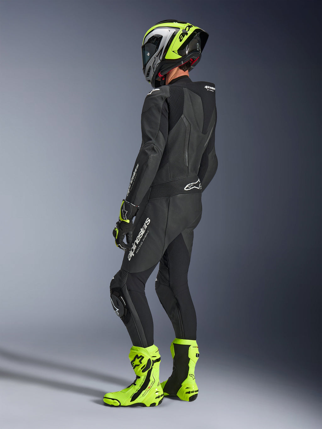 Pilote de moto portant la combinaison Alpinestars GP Force V2 Leather Suit 1-Piece en noir, vue de dos avec logos blancs sur la cuisse et le bas du dos, bosse aérodynamique, associée à un casque jaune fluo et noir, gants et bottes de course assortis, équipement professionnel pour la piste et le sport.