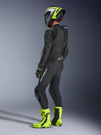 Pilote de moto portant la combinaison Alpinestars GP Force V2 Leather Suit 1-Piece en noir, vue de dos avec logos blancs sur la cuisse et le bas du dos, bosse aérodynamique, associée à un casque jaune fluo et noir, gants et bottes de course assortis, équipement professionnel pour la piste et le sport.