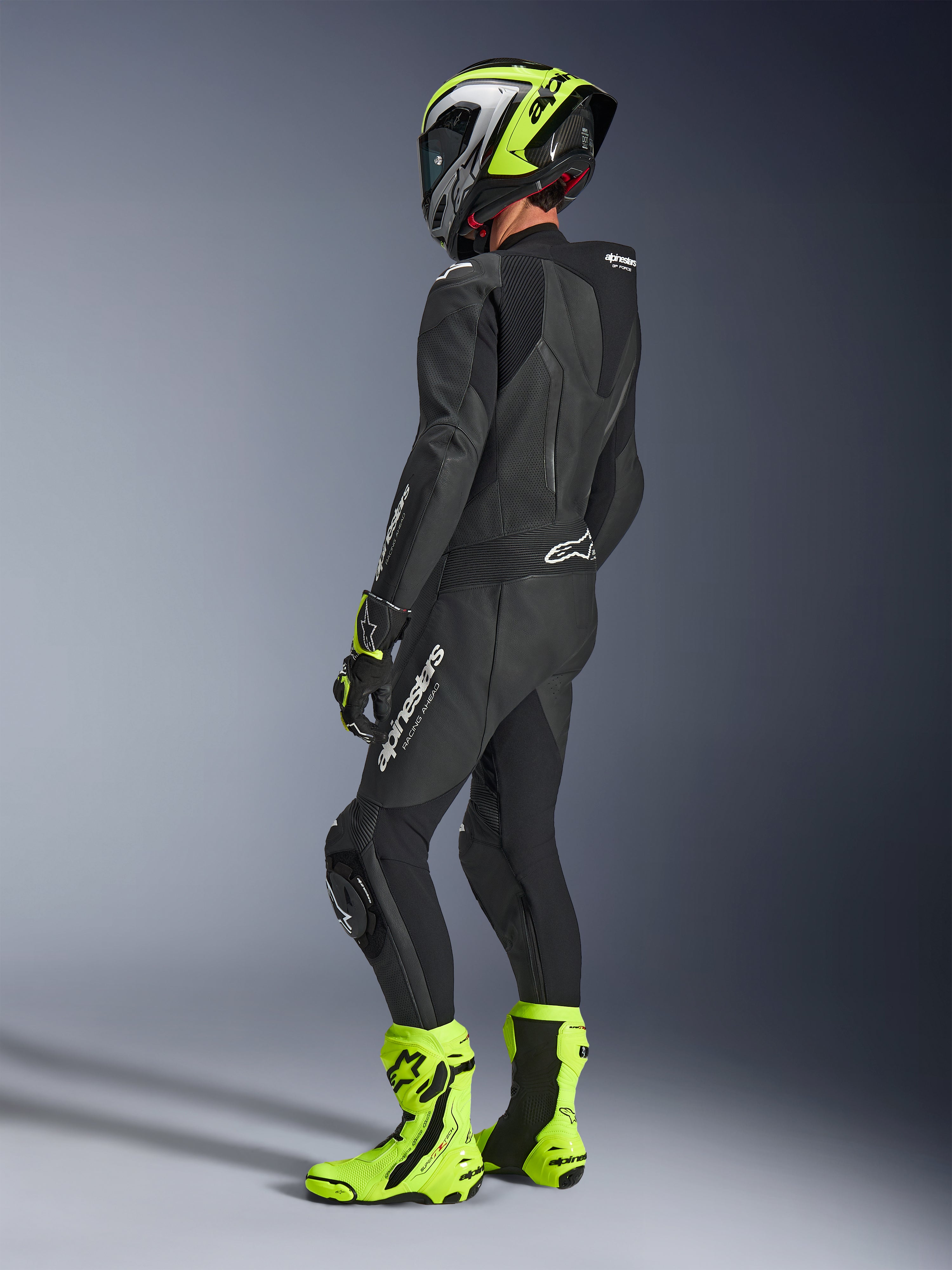 Pilote de moto portant la combinaison Alpinestars GP Force V2 Leather Suit 1-Piece en noir, vue de dos avec logos blancs sur la cuisse et le bas du dos, bosse aérodynamique, associée à un casque jaune fluo et noir, gants et bottes de course assortis, équipement professionnel pour la piste et le sport.
