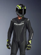 Combinaison en cuir Alpinestars GP Force V2 1 pièce, noire, portée par un pilote avec un casque et des gants noir et jaune fluo, arborant le logo Alpinestars blanc sur la poitrine, des panneaux en cuir perforé pour la ventilation et un design professionnel prêt pour la course.