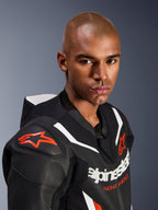 Portrait en gros-plan d'un mannequin portant la combinaison en cuir Alpinestars GP Force V2, 1 pièce, noir, blanc et rouge fluo, avec des panneaux en cuir perforé, des bandes de course blanches et des logos Alpinestars rouges sur l'épaule et la poitrine.