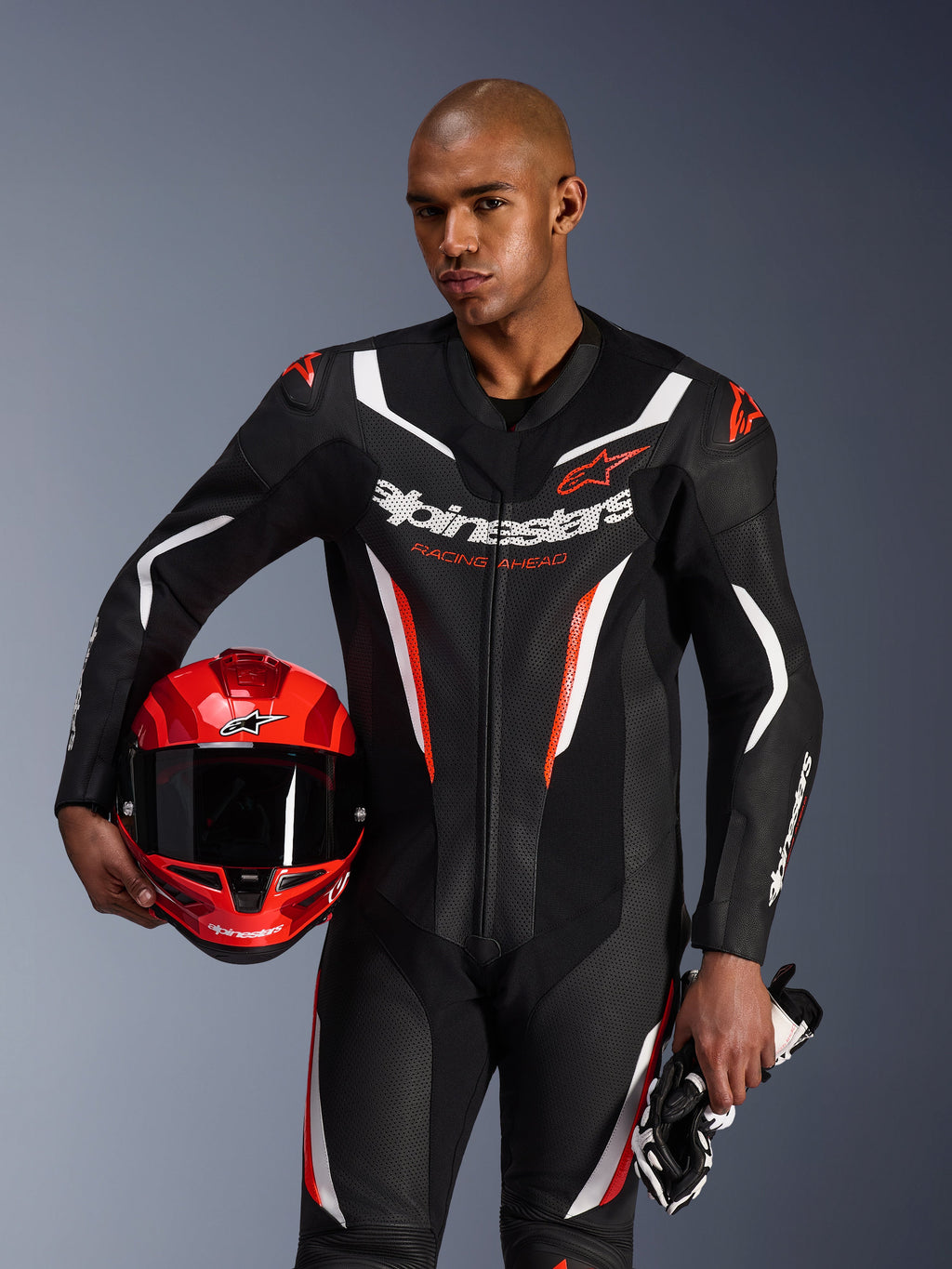 Alpinestars GP Force V2 Leather Suit, combinaison de moto 1 pièce, noir, blanc et rouge fluo, portée par un mannequin tenant un casque et des gants rouges, design racing épuré avec cuir perforé et logos distinctifs