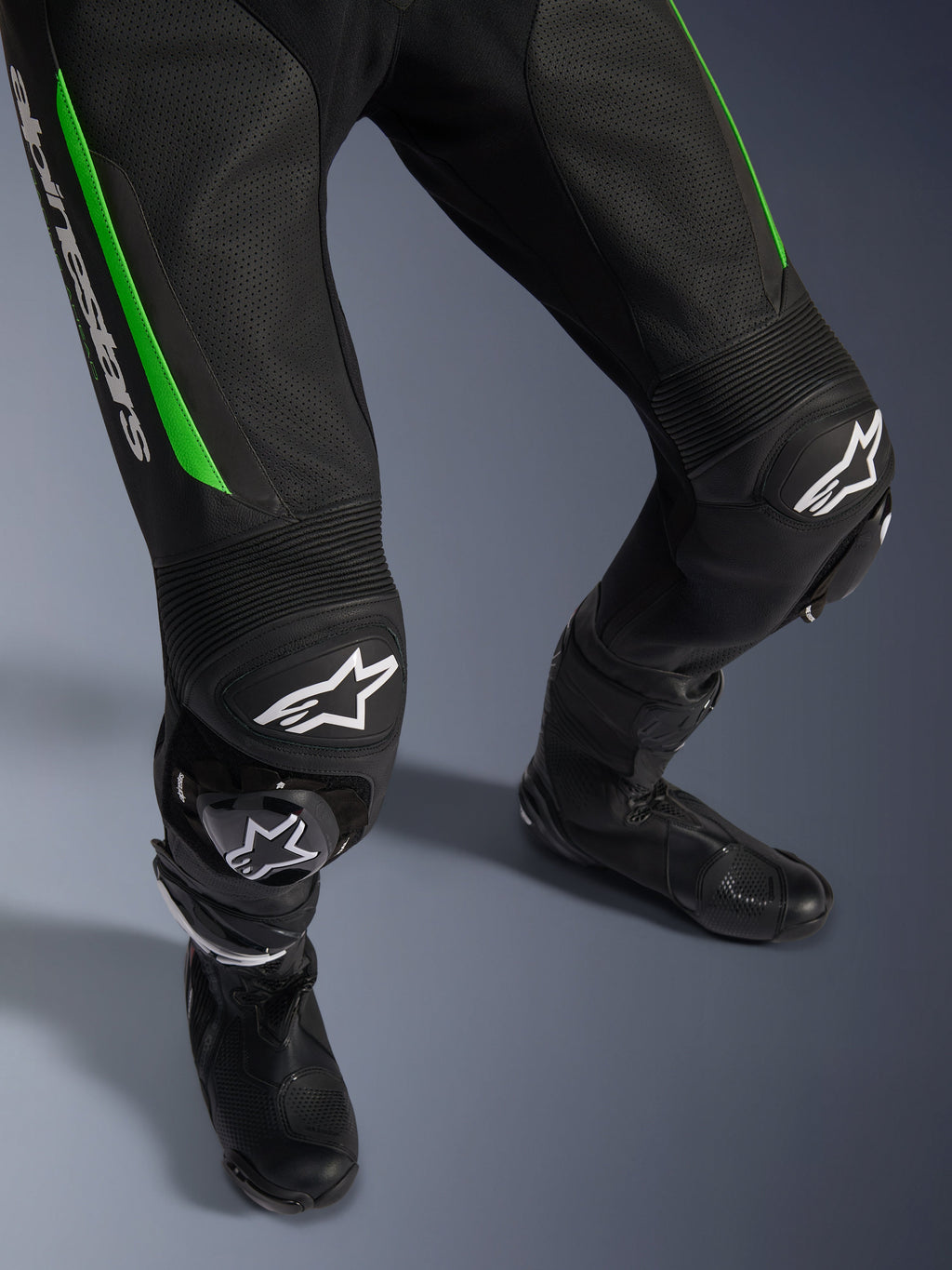 Alpinestars GP Force V2 Leather Suit - 1Piece, close-up of legs in Black and Green, featuring perforated leather construction, accordion stretch panels at the knees, white Alpinestars branding, and integrated knee sliders, worn with black racing boots